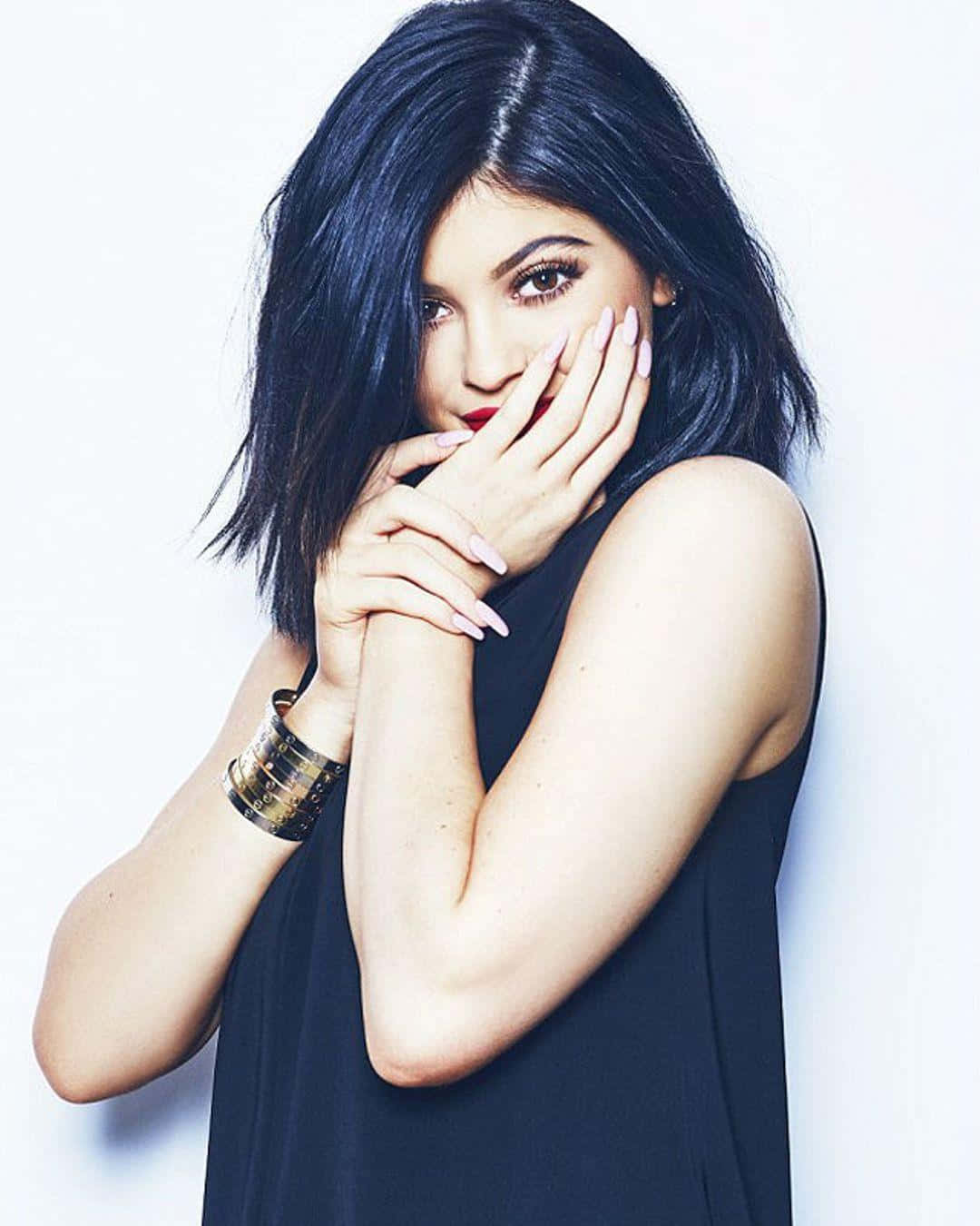 Kylie Jenner 桌布 壁紙 — 一位深色頭髮和紅唇的女性在白色背景前擺姿勢，雙手靠近臉部，身穿黑色上衣和金色手鐲。
