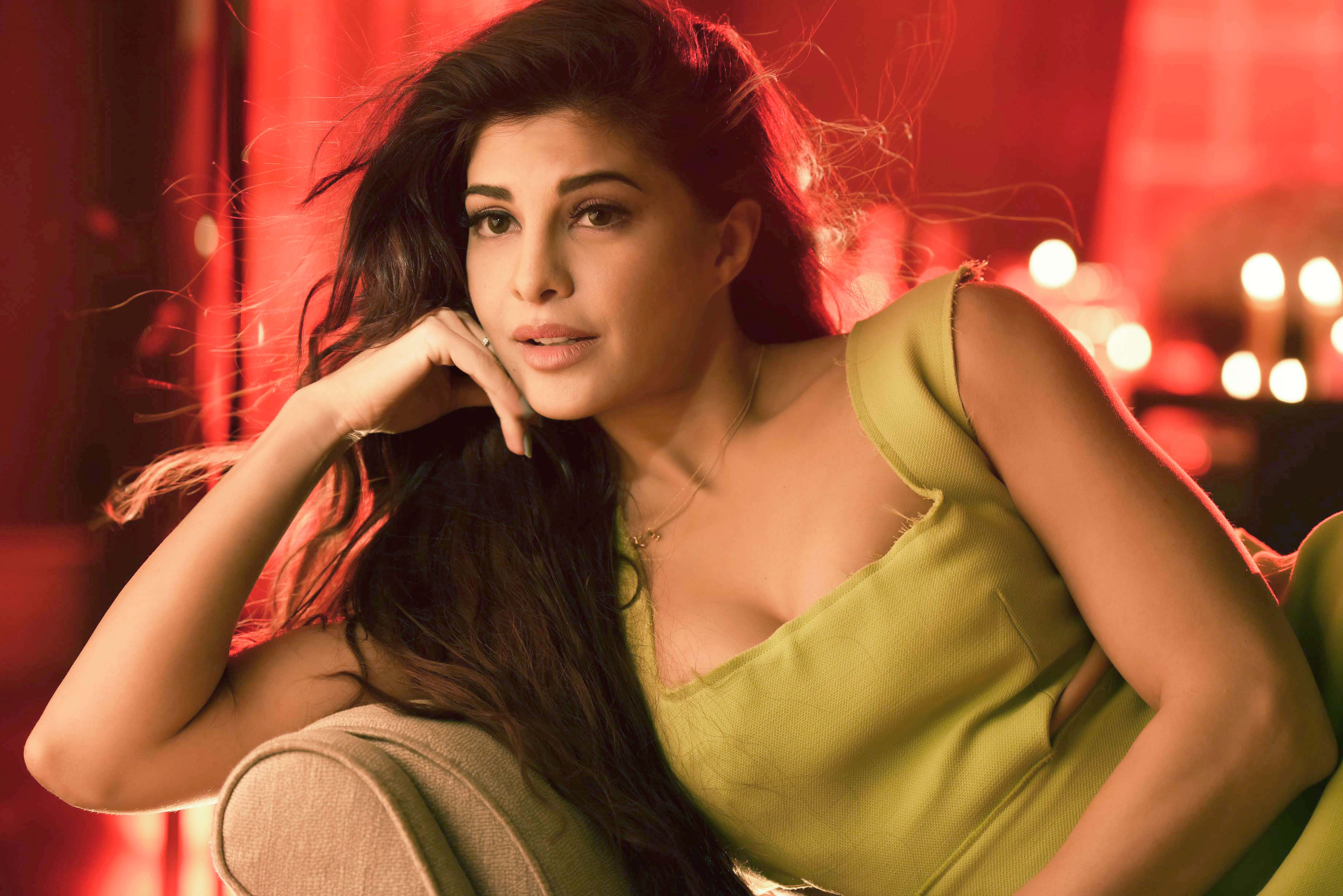 Jacqueline Fernandez 桌布 壁紙 — 一位深色頭髮的女性，在溫暖的紅色燈光背景下，將下巴擱在手上，周圍有柔和發光的燈光。
