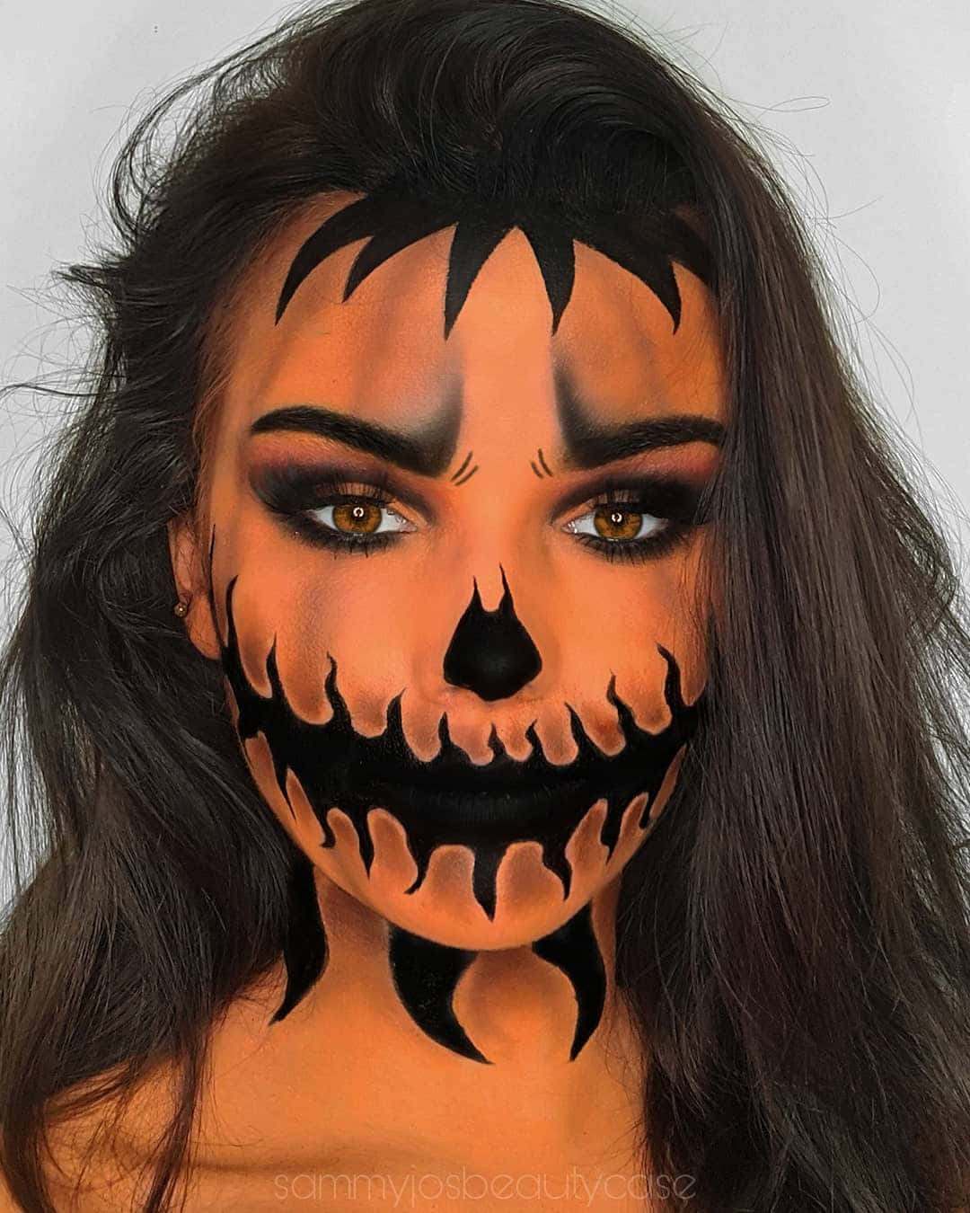Halloween Face Paint 桌布 壁紙 — 一位深色頭髮的女性，臉上畫著戲劇性的萬聖節骷髏妝容，眼睛和嘴巴周圍有橙色和黑色的火焰。