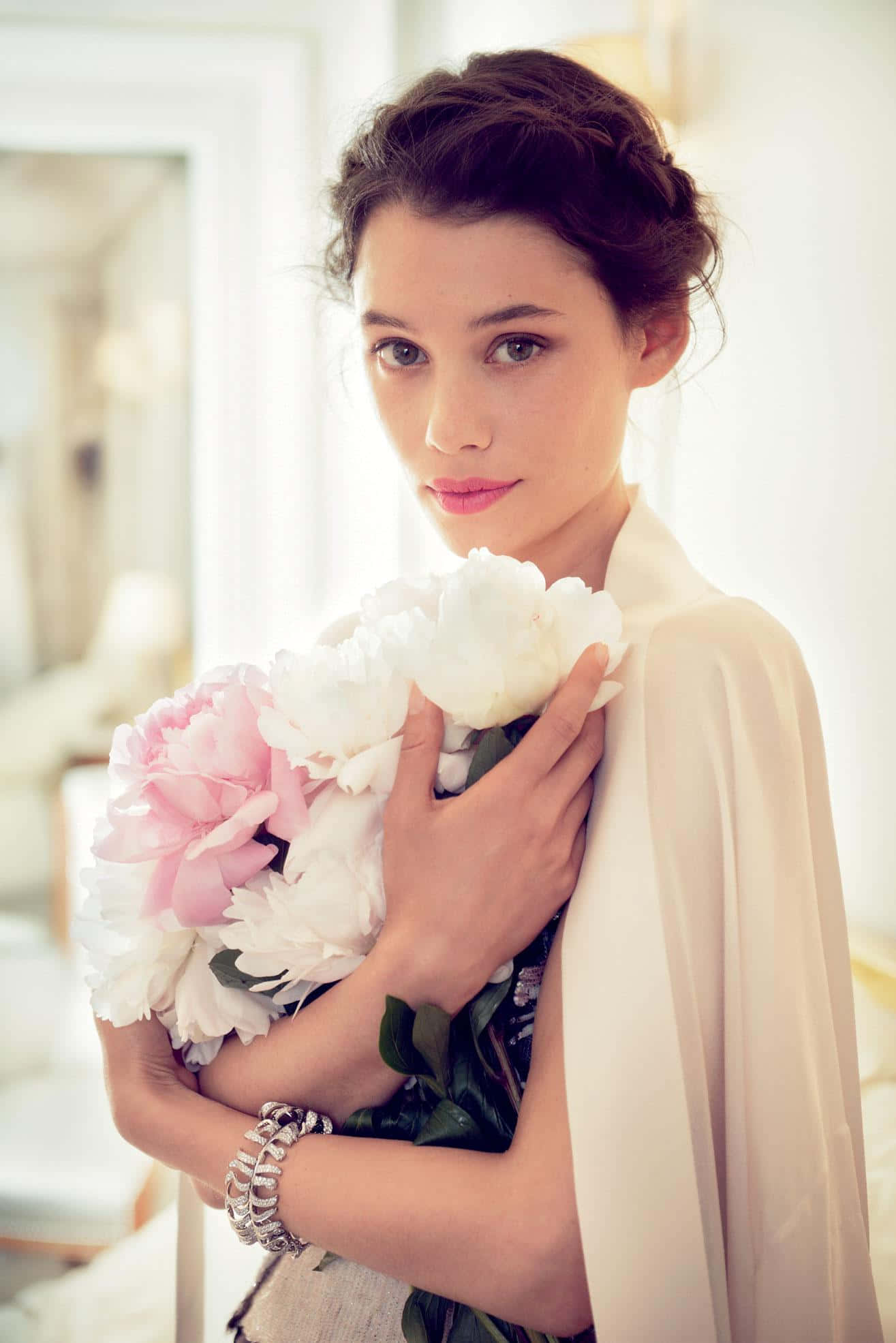 Astrid Berges Frisbey 桌布 壁紙 — 一位深色頭髮的女性在明亮的房間裡，穿著奶油色披肩，手持一束粉紅色和白色的牡丹花。