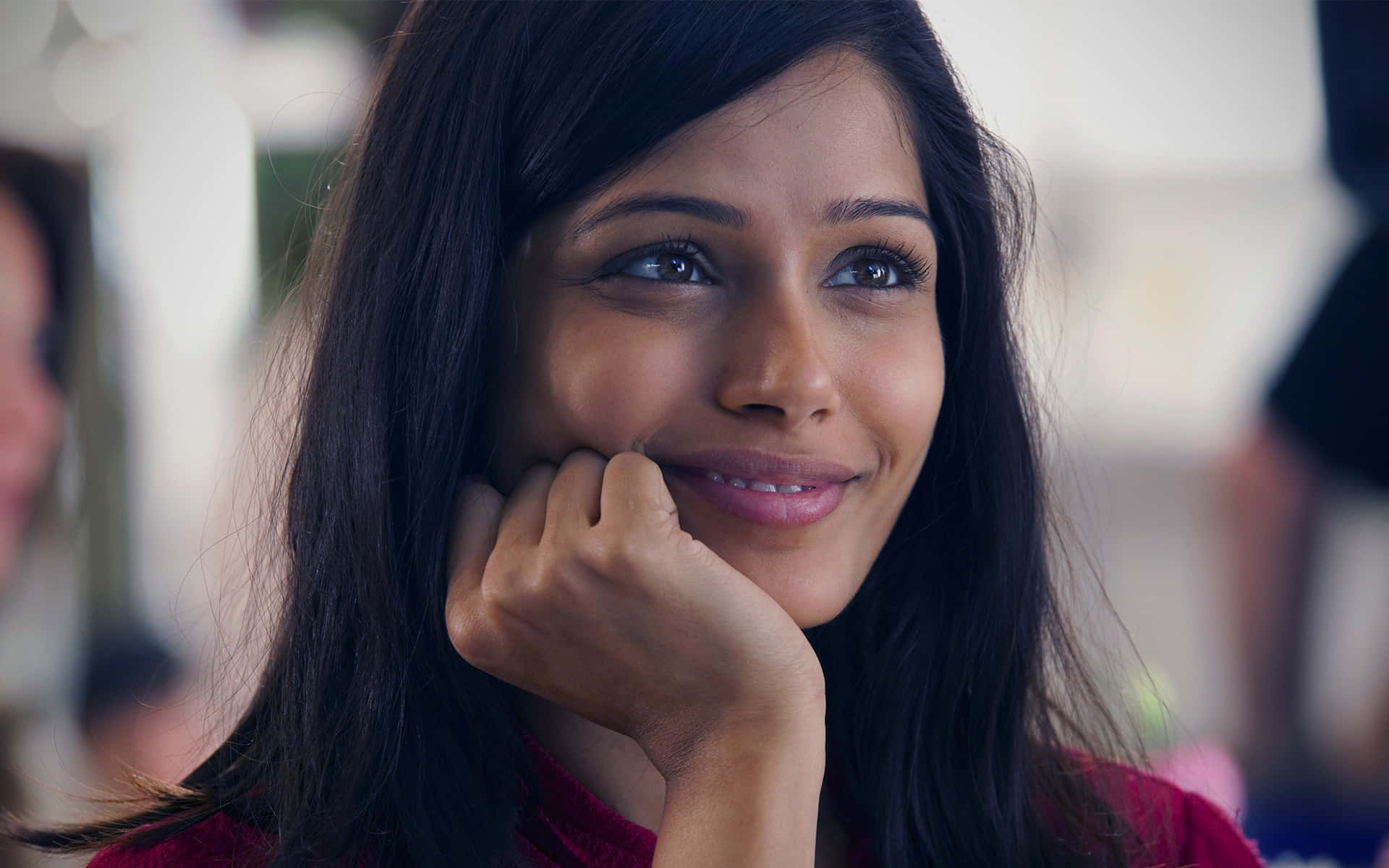 Freida Pinto 桌布 壁紙 — 一位深色頭髮的女性在柔和光線的室內環境中，將下巴靠在手上，溫柔地微笑。