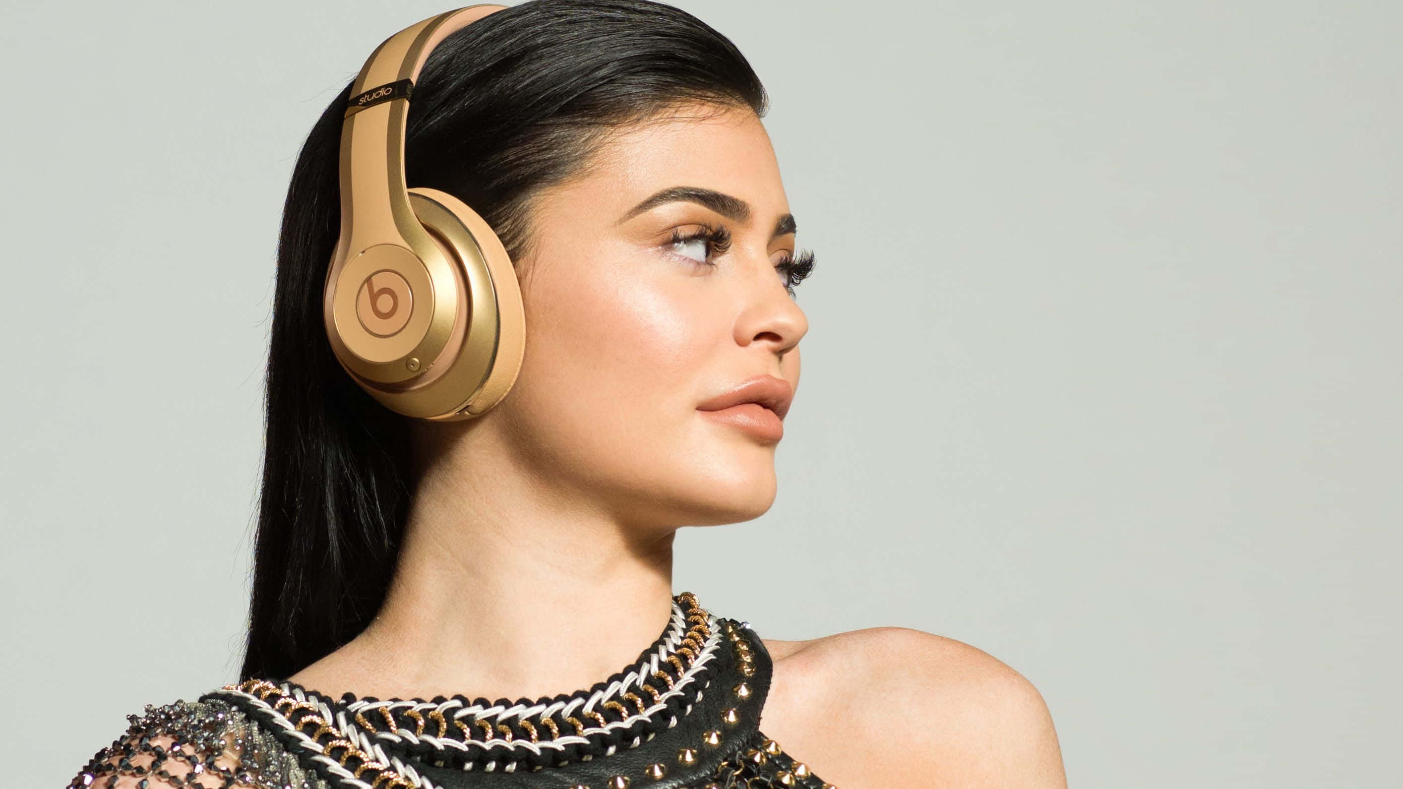 Kylie Jenner 桌布 壁紙 — 一位深色頭髮的女性在純淺灰色背景前佩戴金色 Beats 耳機。