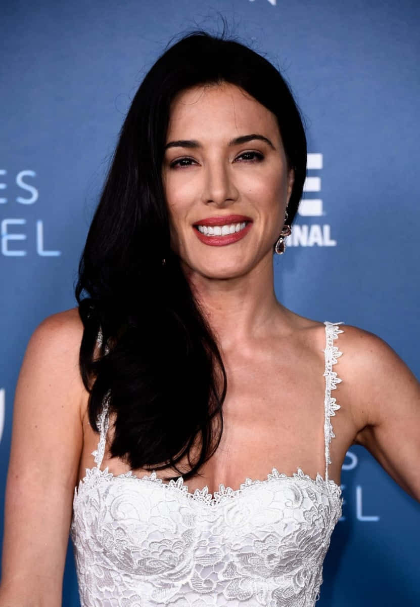 Jaime Murray 桌布 壁紙 — 一位深色頭髮的女性在藍色活動背景前穿著白色蕾絲連衣裙溫暖地微笑。