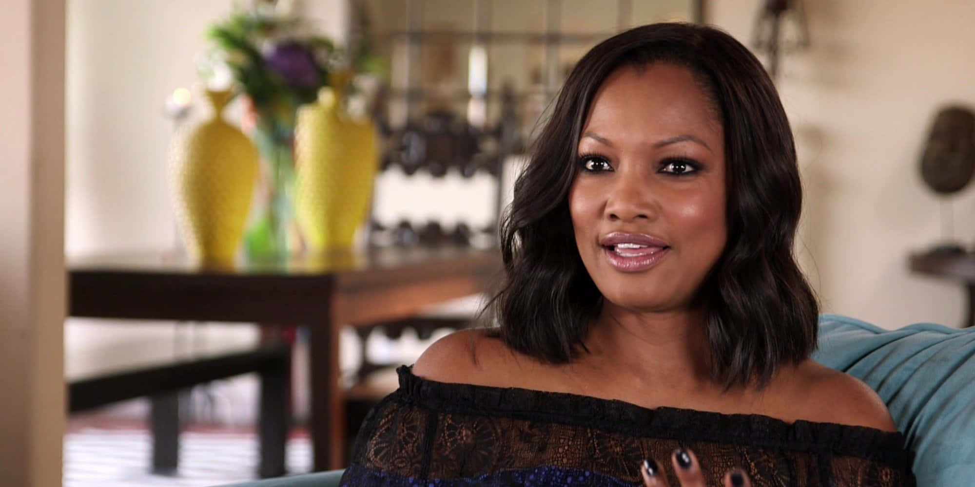 Garcelle Beauvais 桌布 壁紙 — 一位深色頭髮的女性坐在藍色沙發上，在柔和光線的室內環境中溫暖地微笑。