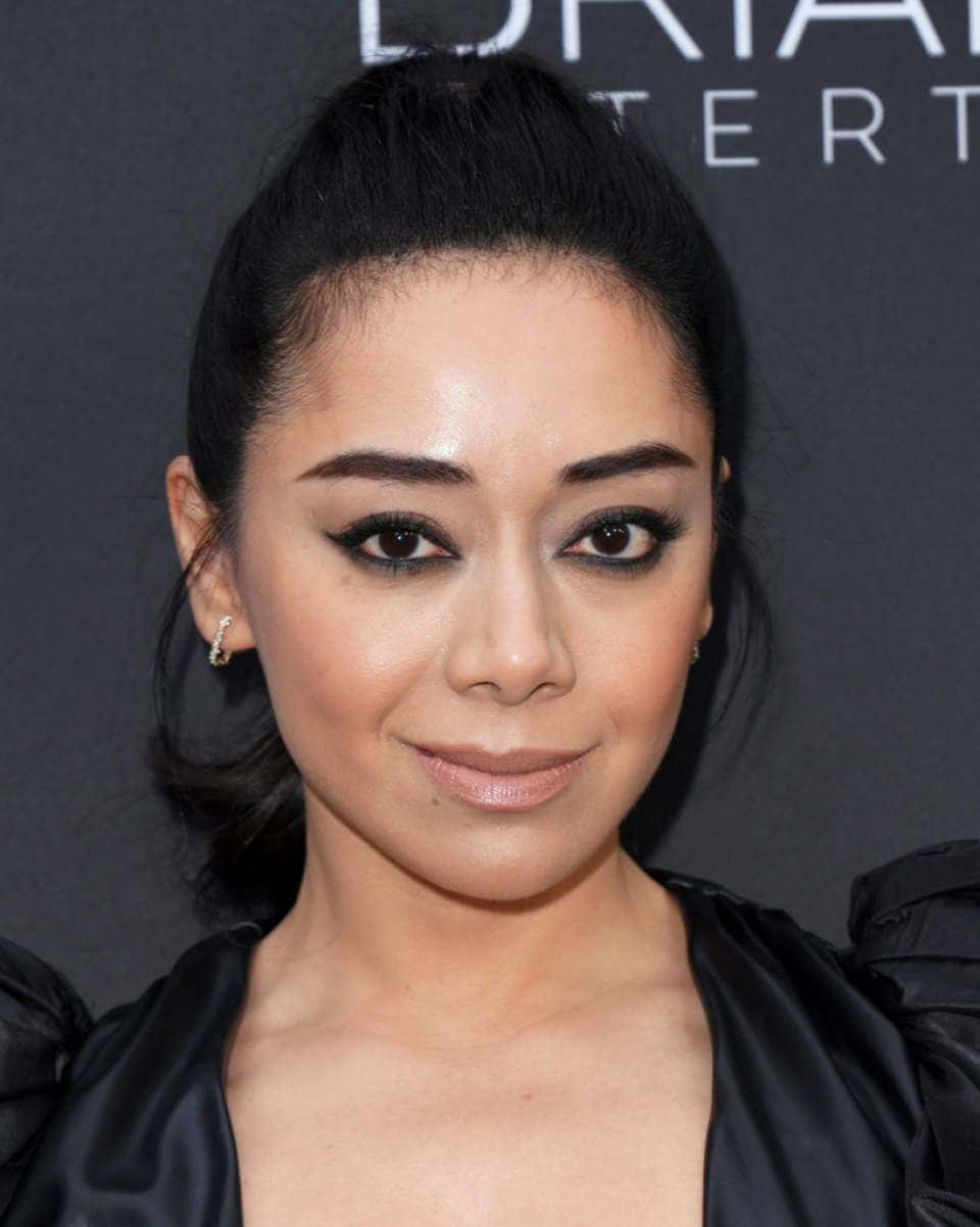 Aimee Garcia 桌布 壁紙 — 一位深色頭髮高馬尾的女性特寫肖像，穿著黑色緞面服裝和淡妝，背景為深色品牌背景。