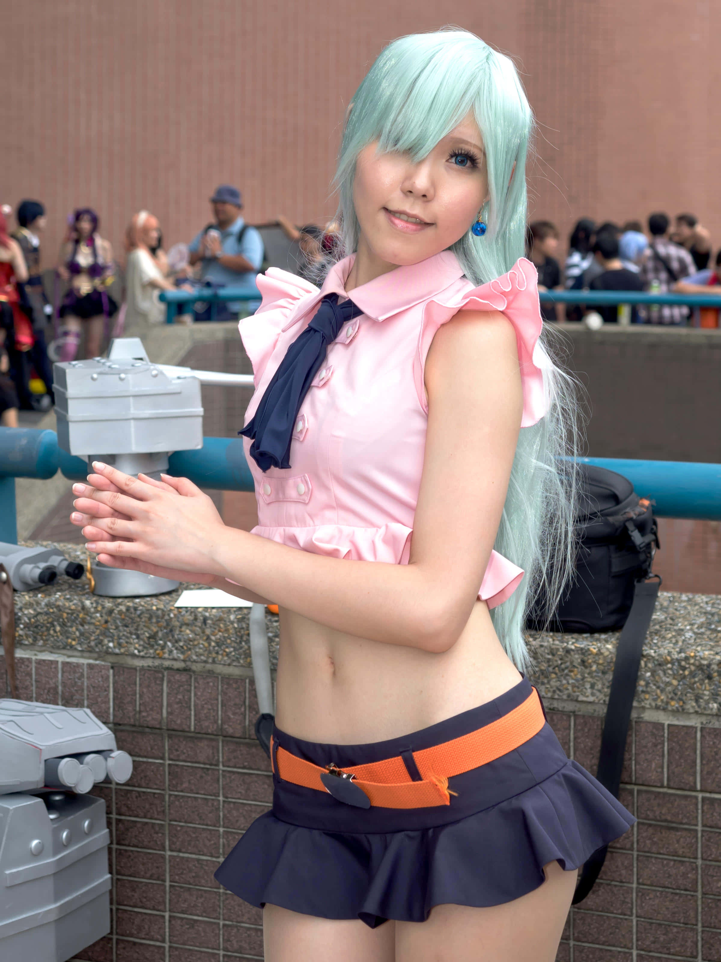 Seven Deadly Sins Elizabeth Liones 桌布 壁紙 — 一位淺藍色頭髮、身穿粉紅色服裝的角色扮演者在會議上自信地擺姿勢，身後有灰色道具和人群。