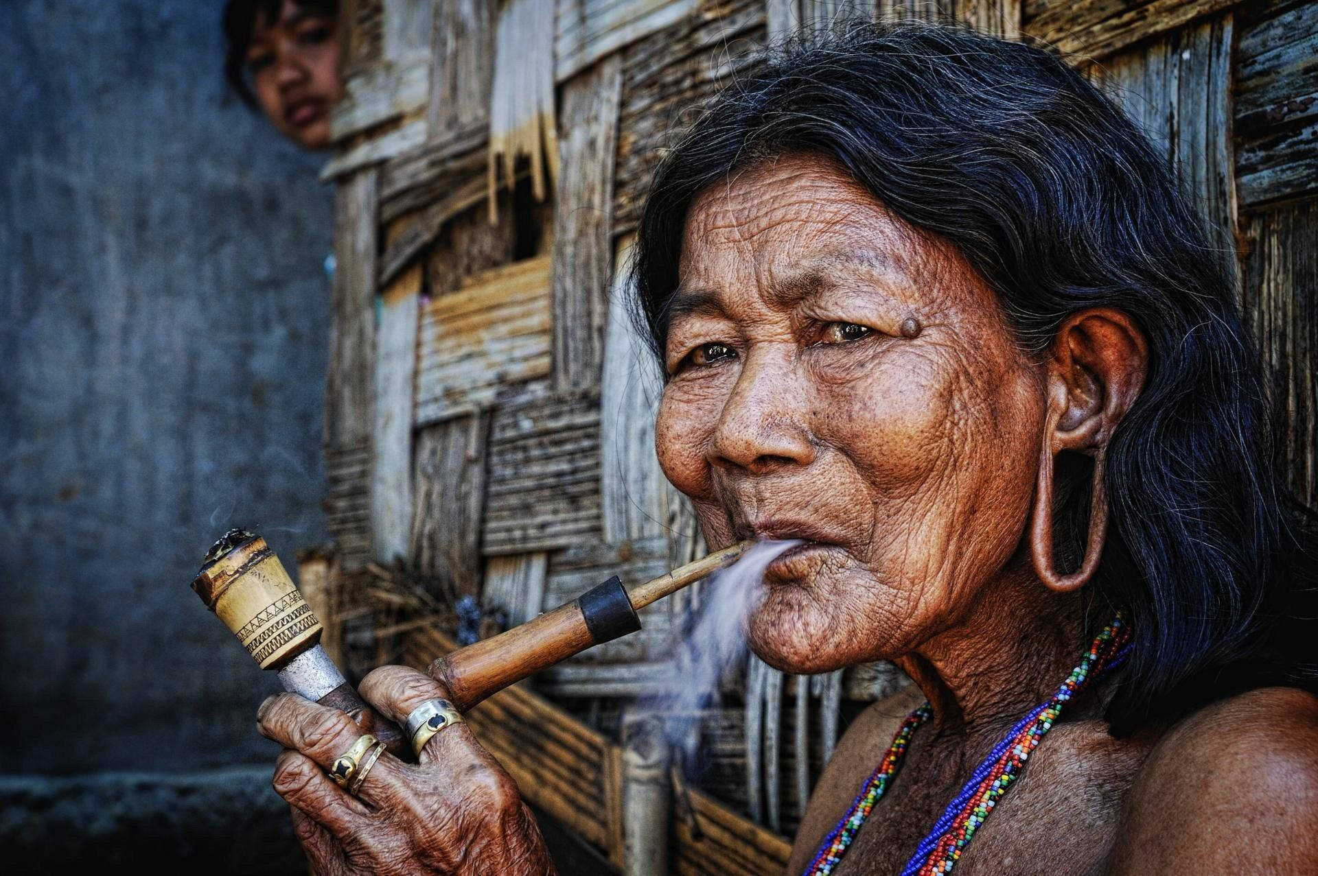 photography 桌布 壁紙 — 一位滿臉皺紋、戴著大耳環的老婦人，站在風化的木屋前抽著竹煙斗。