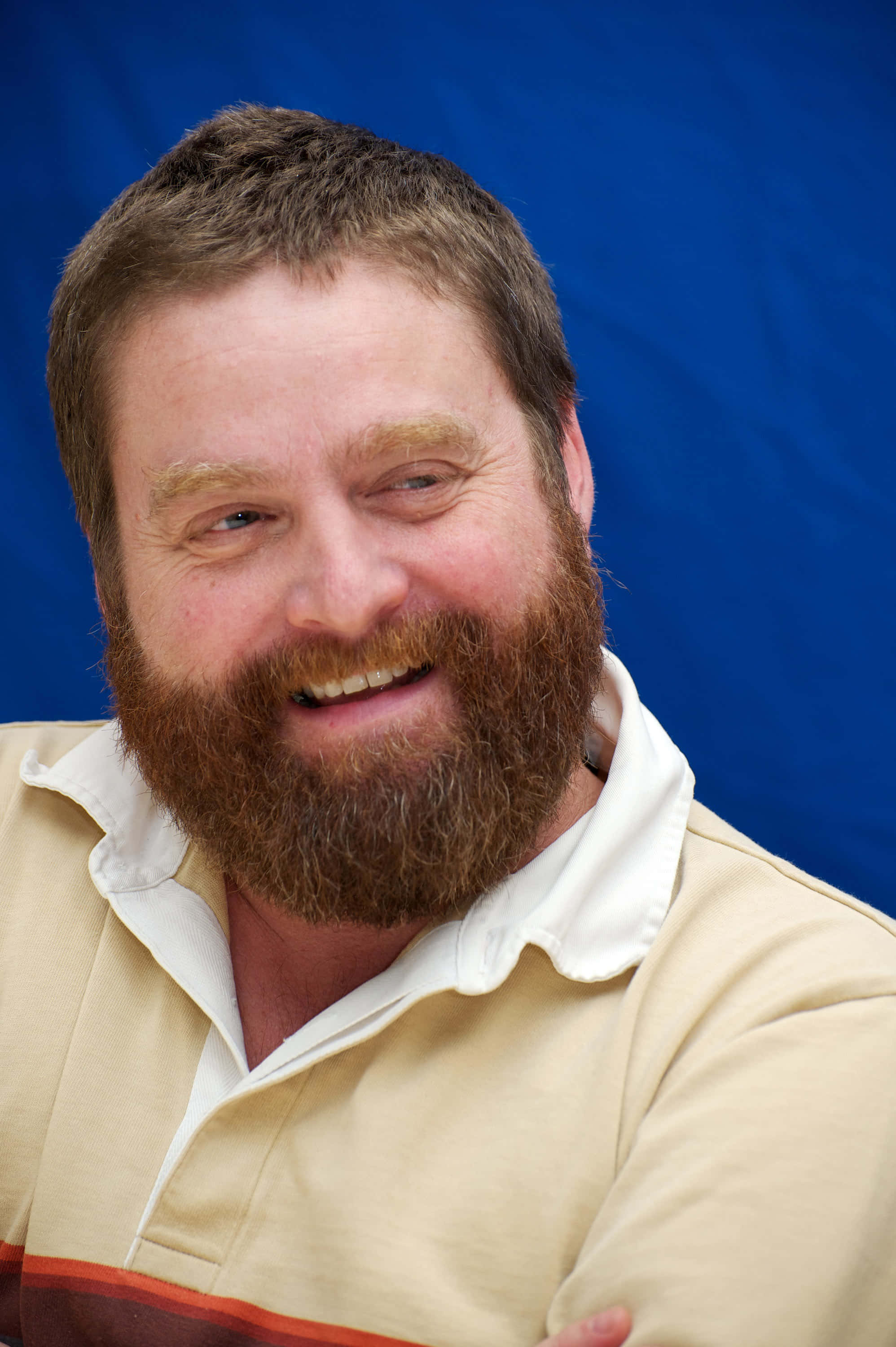 Zach Galifianakis 桌布 壁紙 — 一位滿臉紅棕色鬍鬚的開朗男子，在純藍色背景前溫暖地微笑。