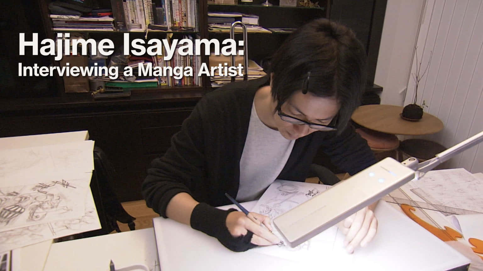 Isayama Hajime 桌布 壁紙 — 一位漫畫家在明亮的燈光下，於雜亂的書桌前專注地素描，身處舒適的家庭工作室。