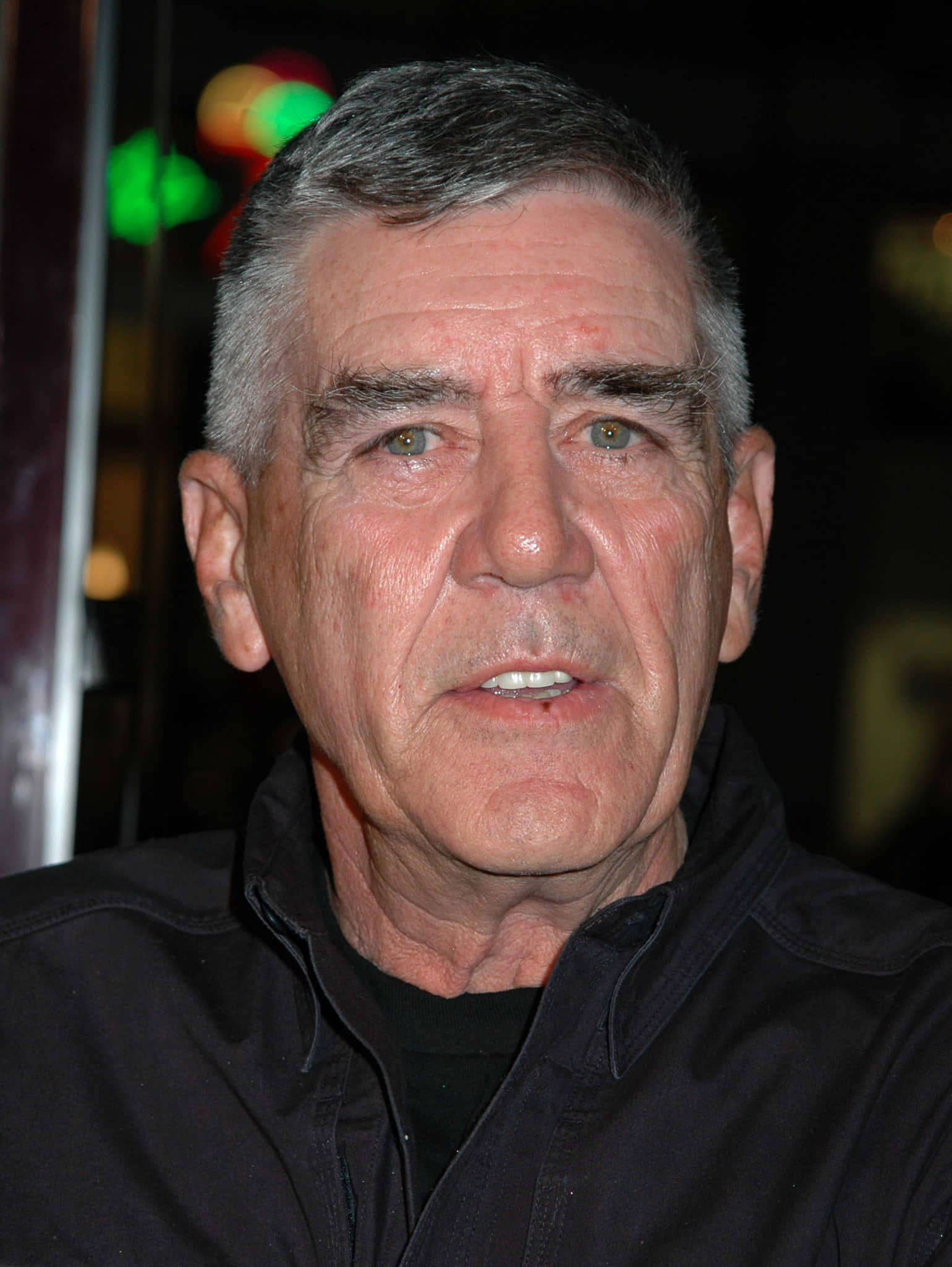 R. Lee Ermey 桌布 壁紙 — 一位灰白頭髮、擁有強烈綠色眼睛的年長男性穿著黑色外套的特寫肖像，背景是模糊的夜晚和彩色散景燈光。