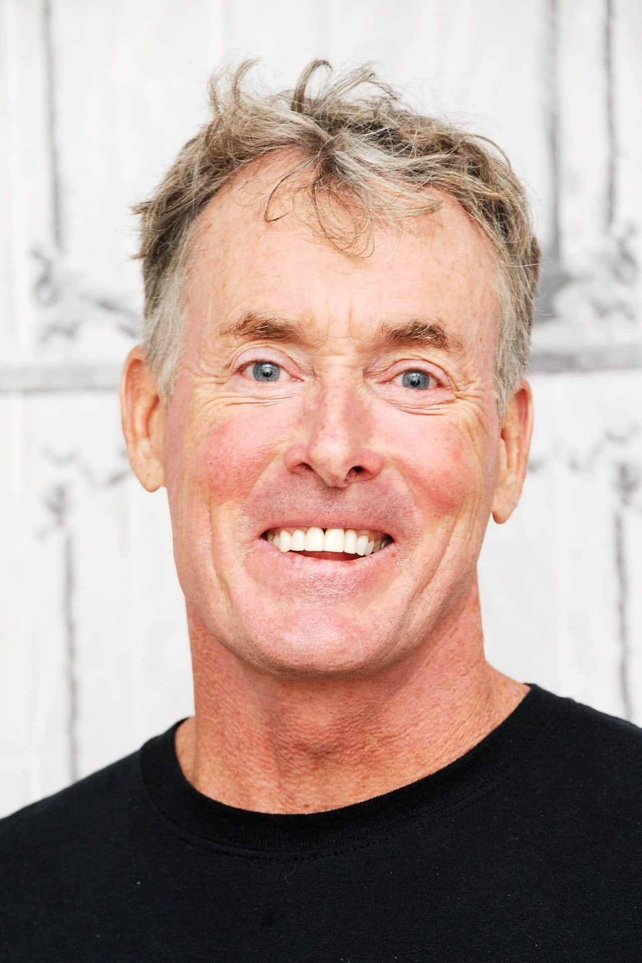 John C. Mcginley 桌布 壁紙 — 一位灰白頭髮、明亮藍眼睛的開朗中年男子，在白色木質背景前溫暖地微笑。