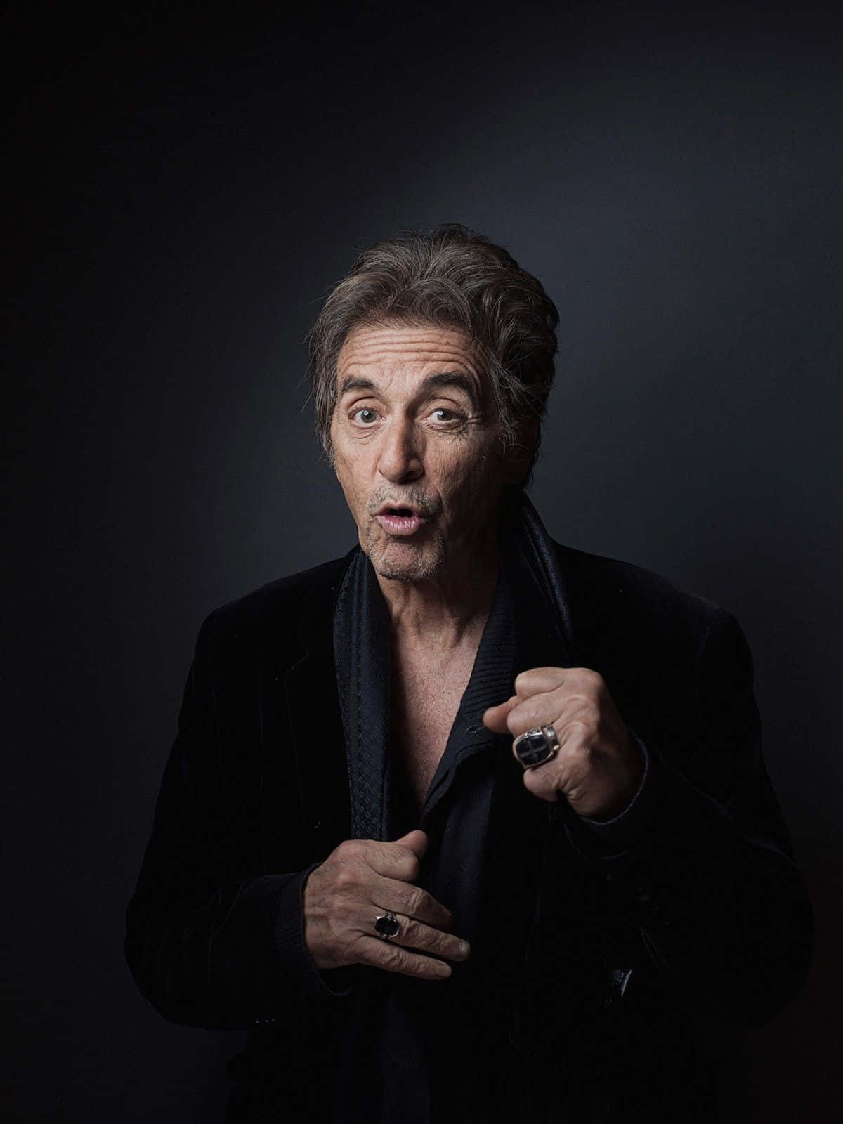 Al Pacino 桌布 壁紙 — 一位灰髮且眼神富有表現力的男士，穿著一件解開釦子的黑色天鵝絨外套，露出深色圍巾和戒指，在深色背景下顯得驚訝。