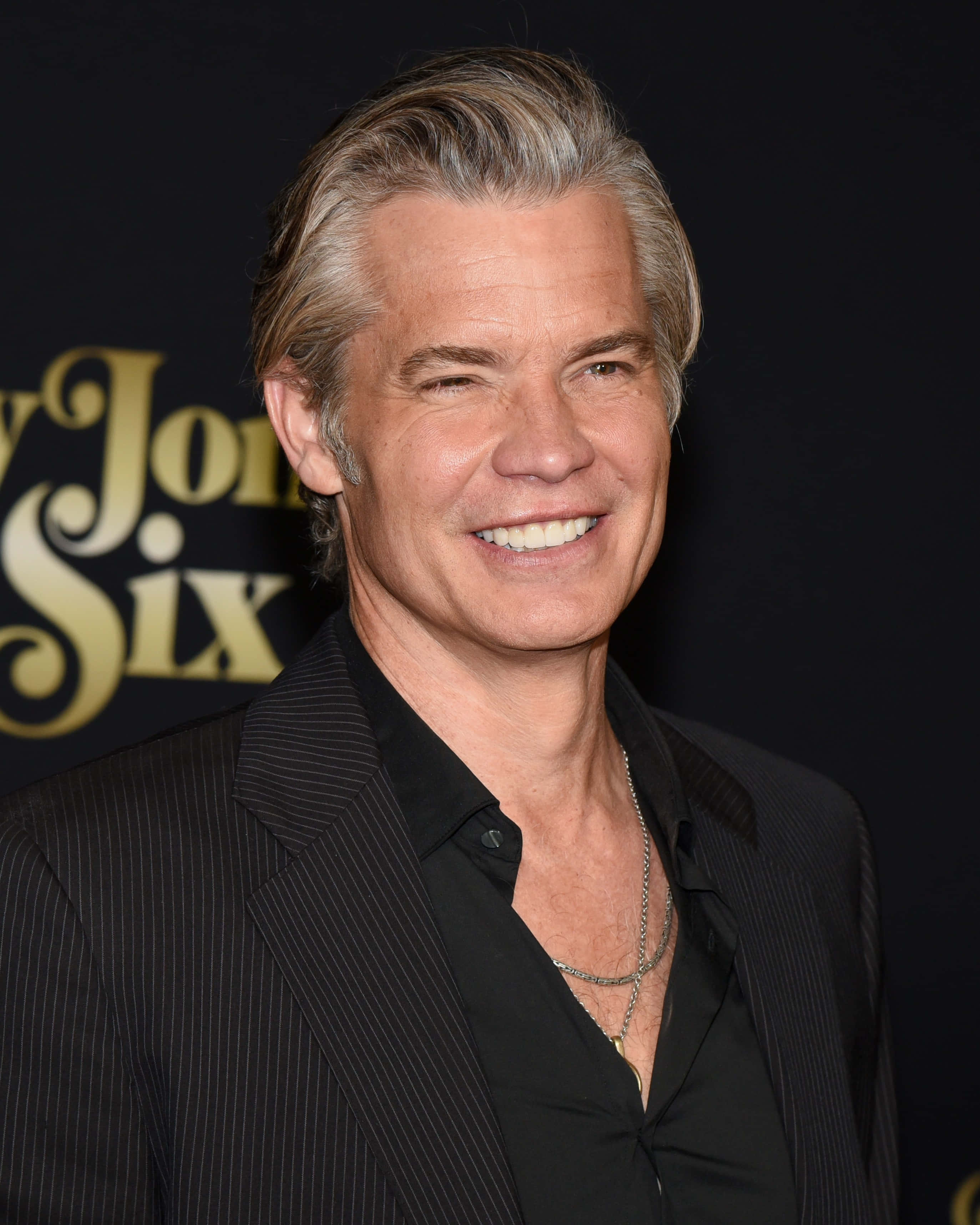 timothy olyphant 桌布 壁紙 — 一位灰髮微笑的男子穿著黑色細條紋西裝，背景為深色並帶有金色文字。