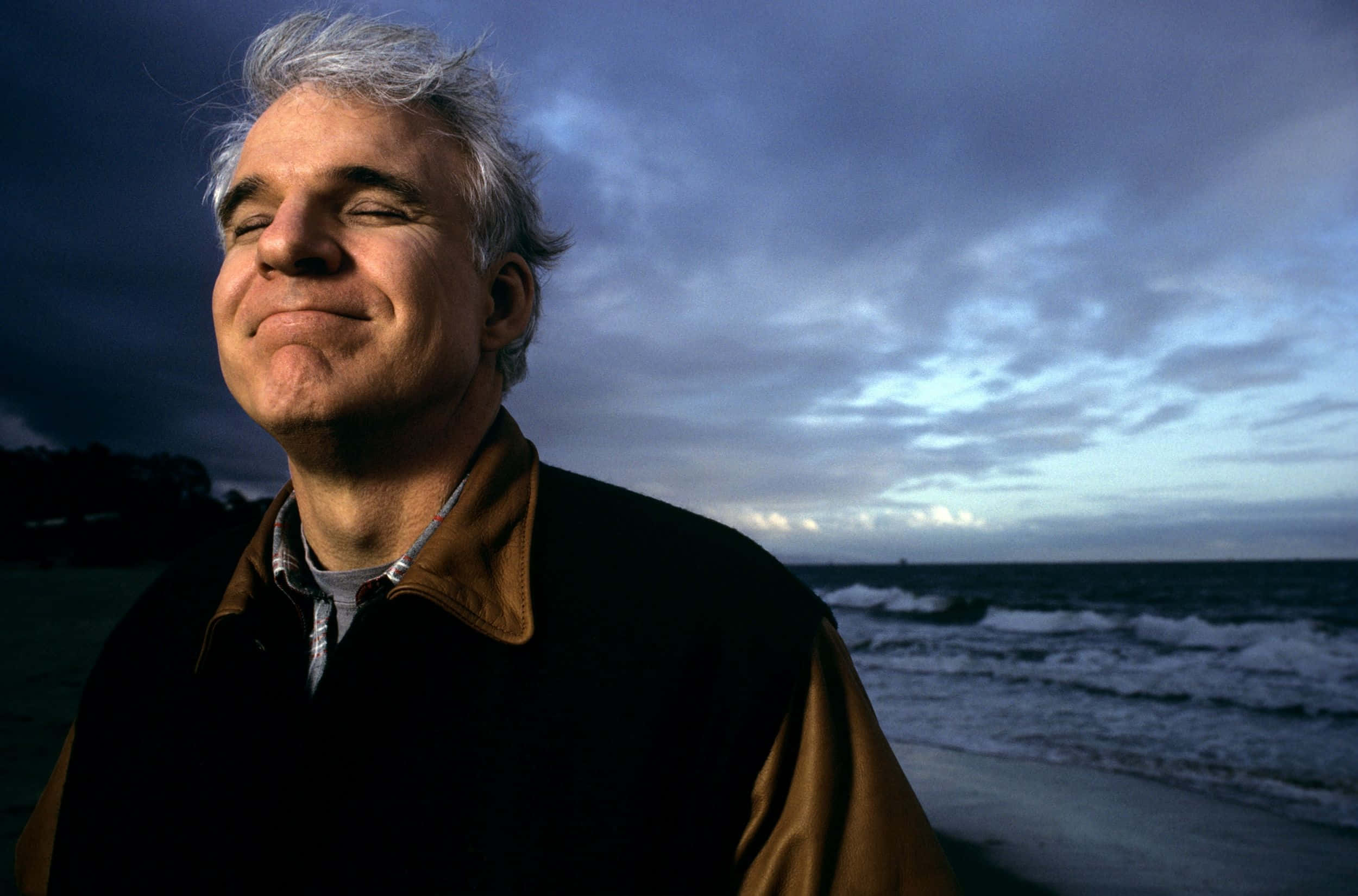 steve martin 桌布 壁紙 — 一位灰髮男子在陰沉暮色海洋背景下，伴隨著烏雲和拍岸海浪，平靜地微笑。