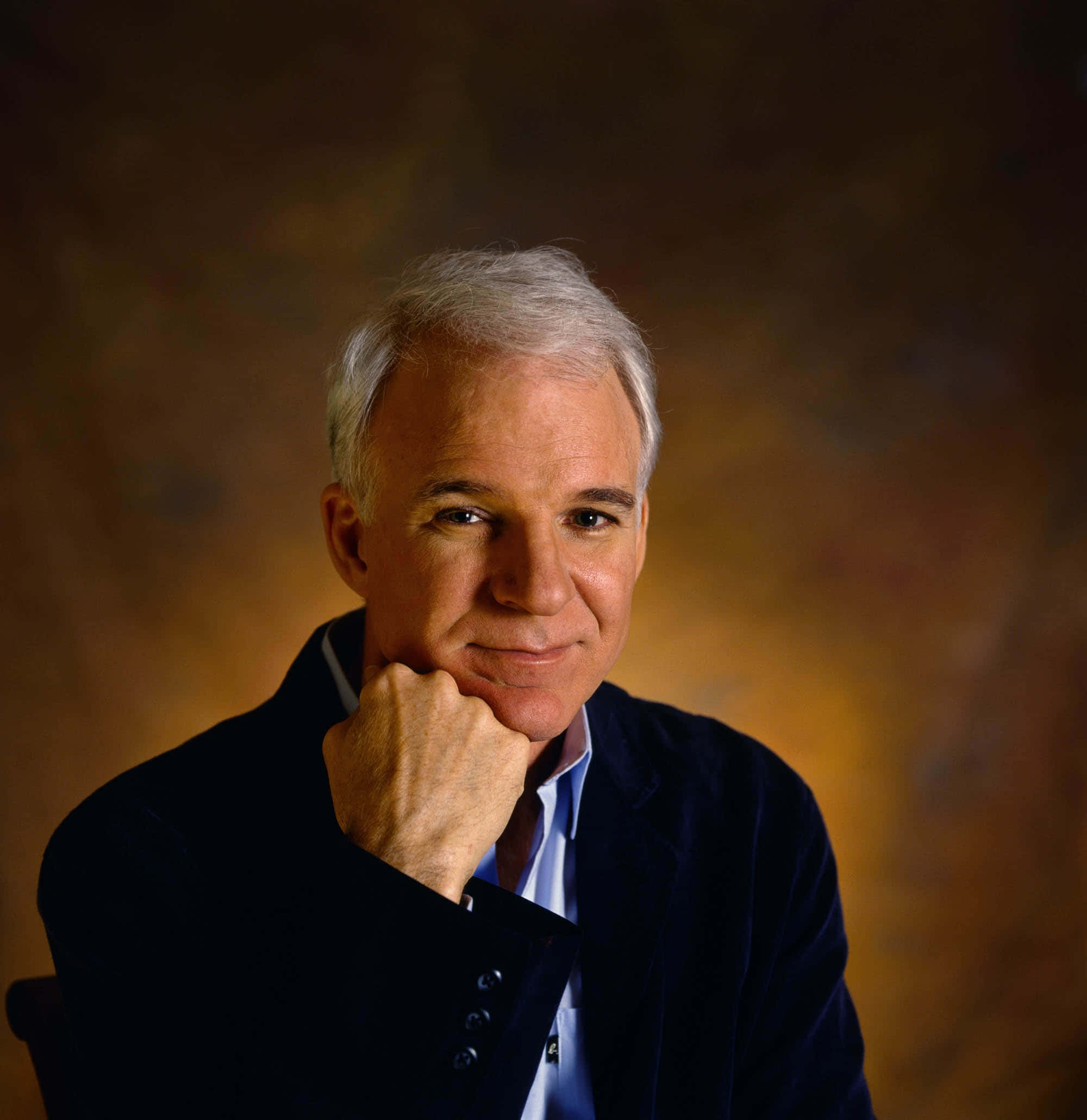 steve martin 桌布 壁紙 — 一位灰髮的成熟男士，在溫暖的漸變背景前，將下巴擱在手上。