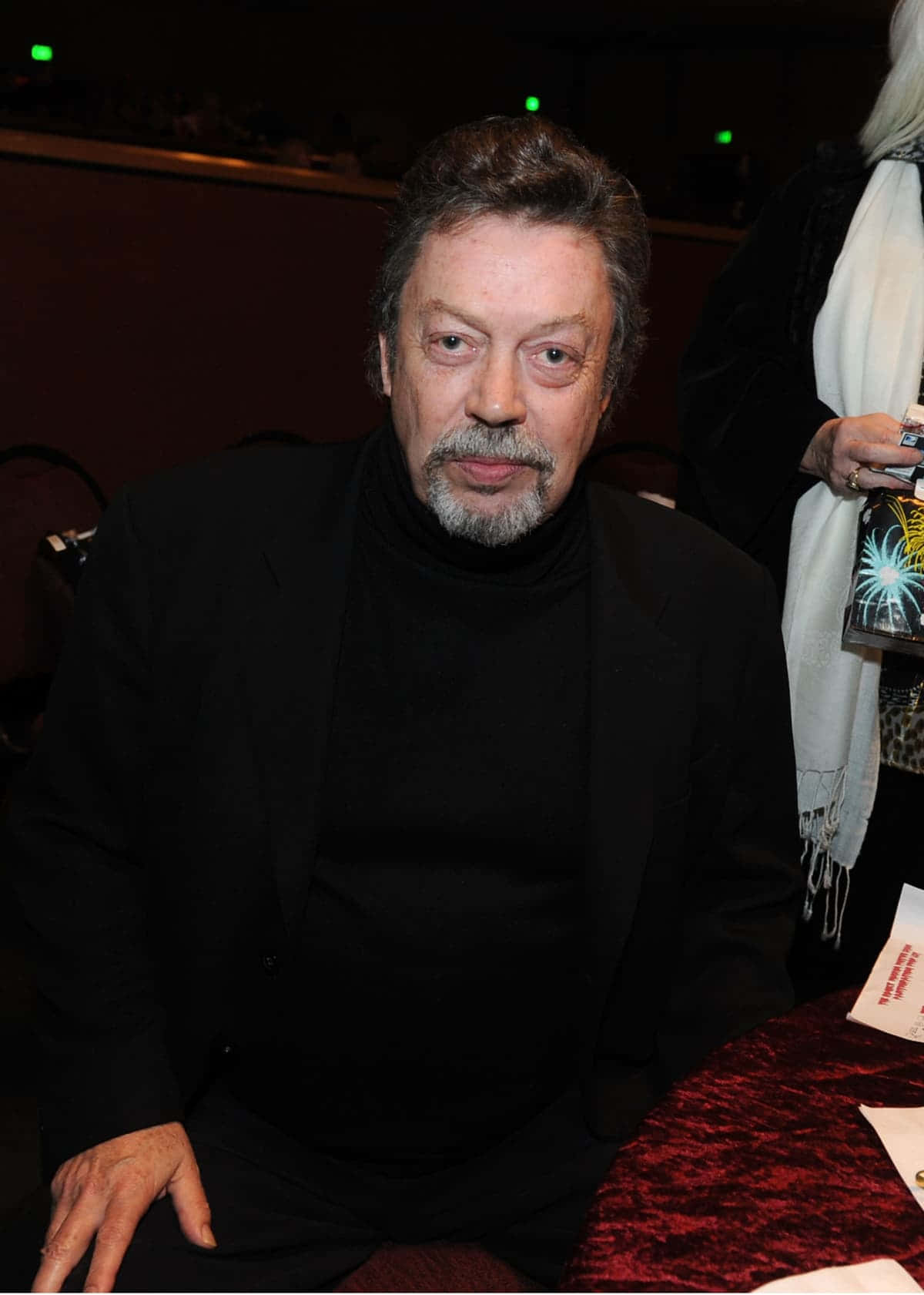 Tim Curry 桌布 壁紙 — 一位灰髮鬍鬚男士坐在燈光昏暗的房間裡，身穿黑色高領毛衣和西裝外套。