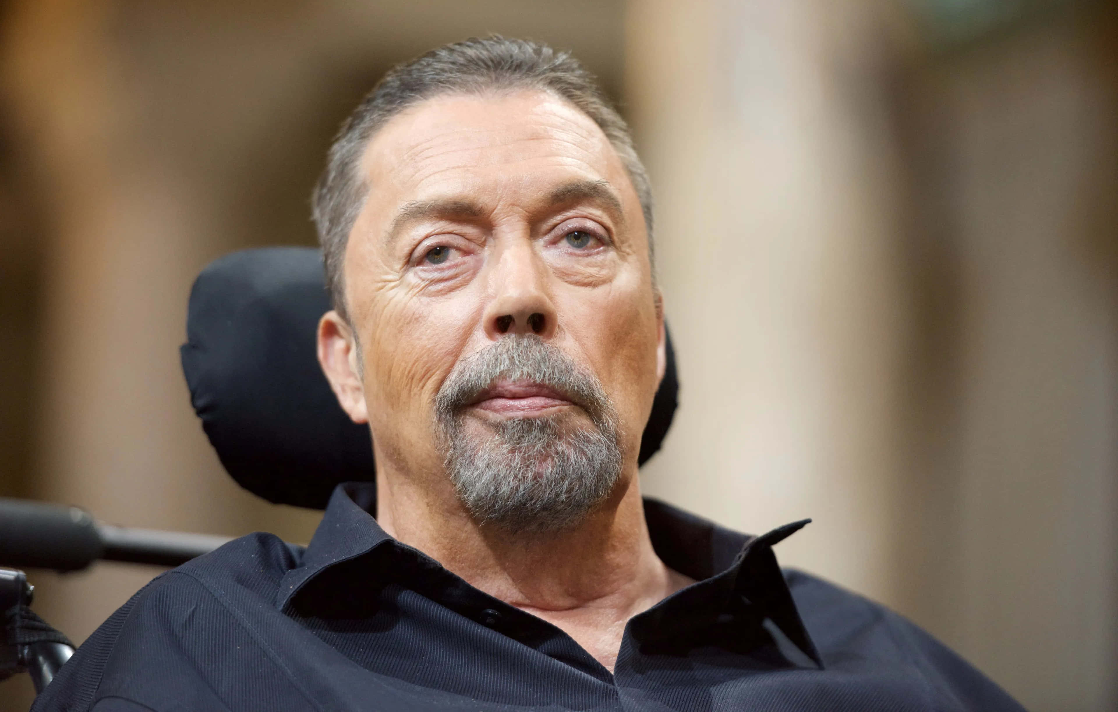 Tim Curry 桌布 壁紙 — 一位灰髮鬍鬚男子坐在輪椅上穿著深色襯衫的特寫肖像。