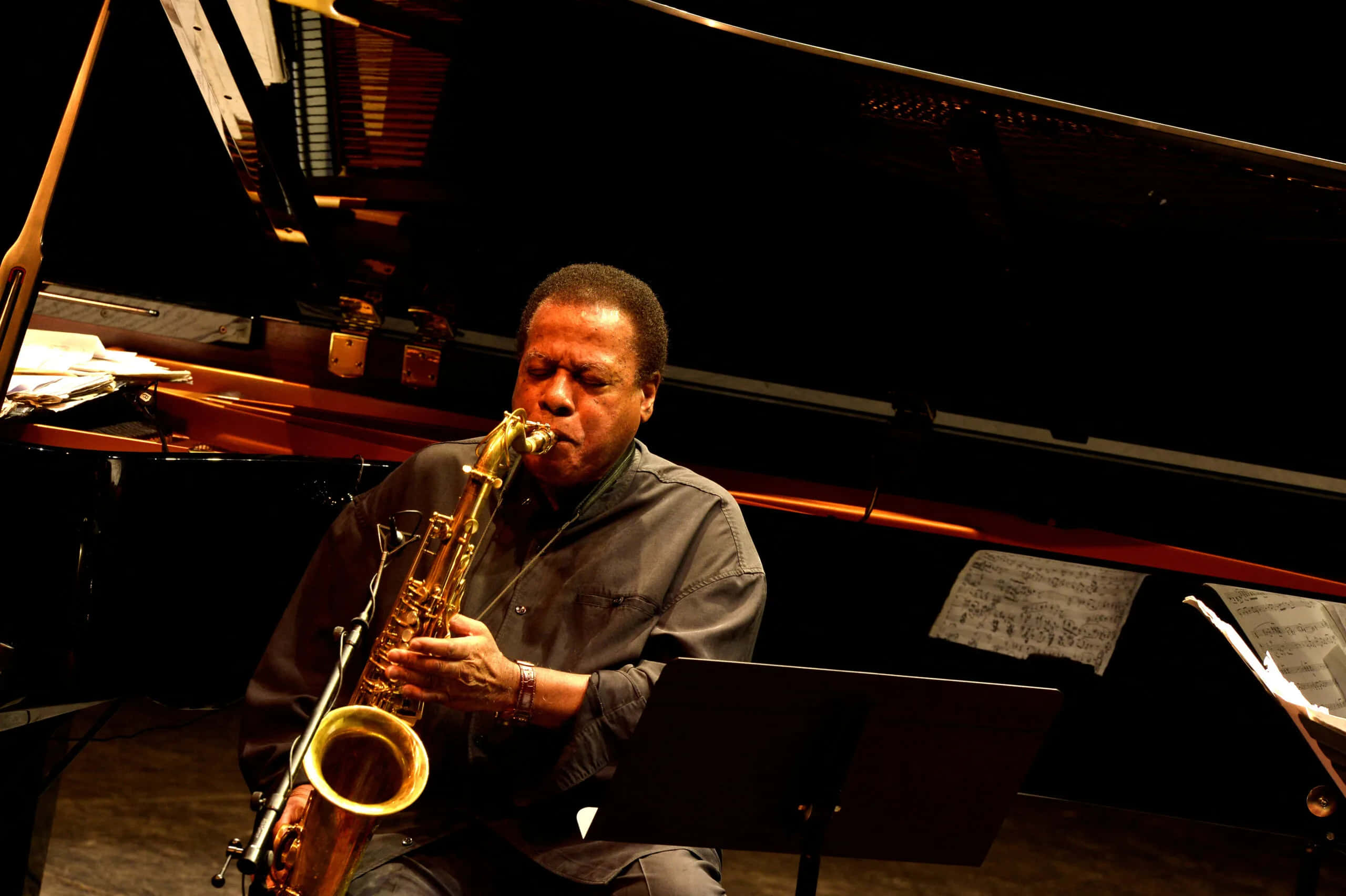 Wayne Shorter 桌布 壁紙 — 一位爵士樂手在溫暖的舞台燈光下，於三角鋼琴旁熱情地吹奏金色色士風。