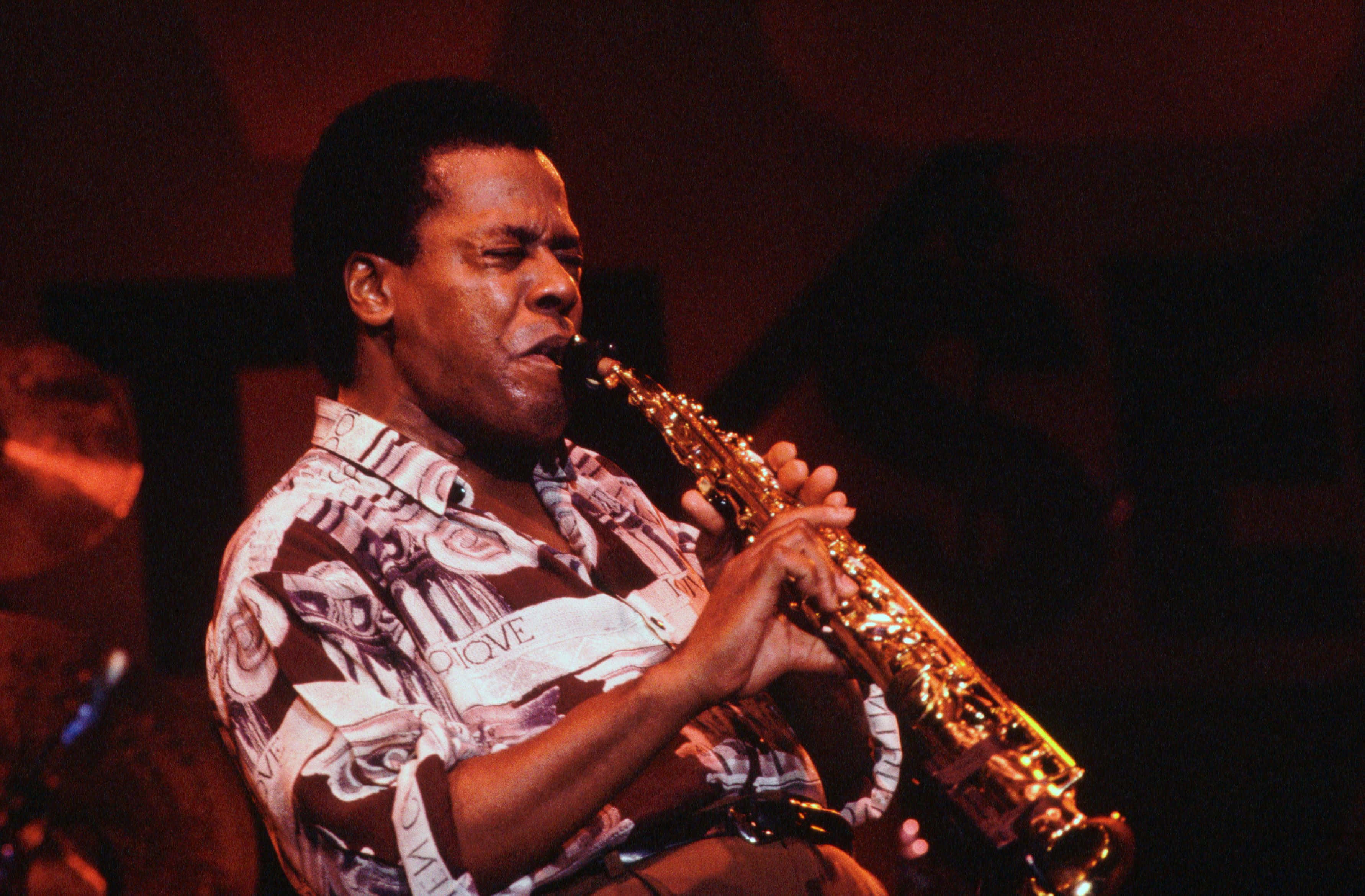 Wayne Shorter 桌布 壁紙 — 一位爵士音樂家在昏暗的俱樂部中，在溫暖的舞台燈光下熱情地吹奏金色薩克斯風。