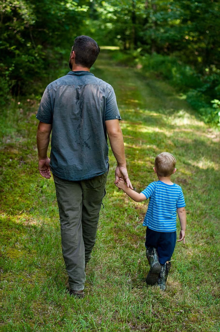 Dad 桌布 壁紙 — 一位父親和年幼的兒子手牽手走在陽光明媚的森林小徑上，周圍是茂密的綠樹。
