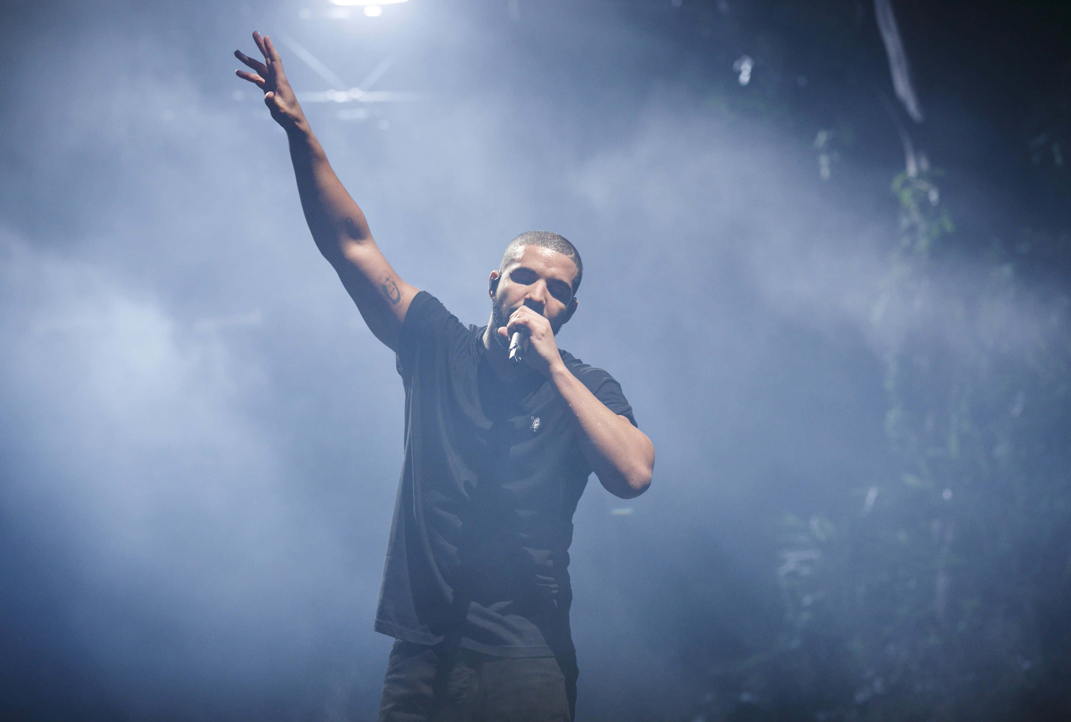Drake 桌布 壁紙 — 一位男性表演者在煙霧彌漫的舞台上，在戲劇性的藍色燈光下，舉起一隻手臂對著麥克風唱歌。