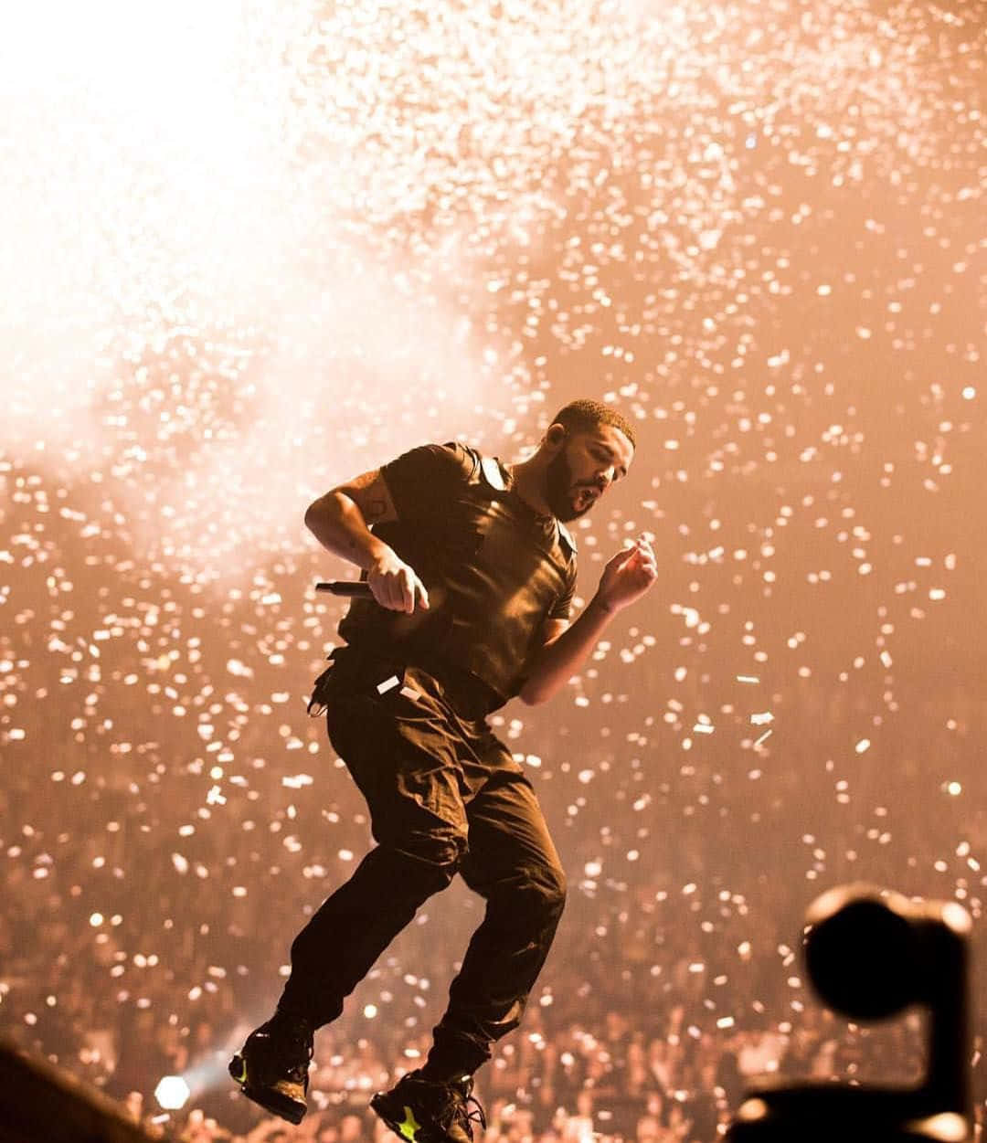 Drake 桌布 壁紙 — 一位男性表演者在舞台上凌空躍起，金色紙屑在溫暖的舞台燈光下在他周圍爆炸。