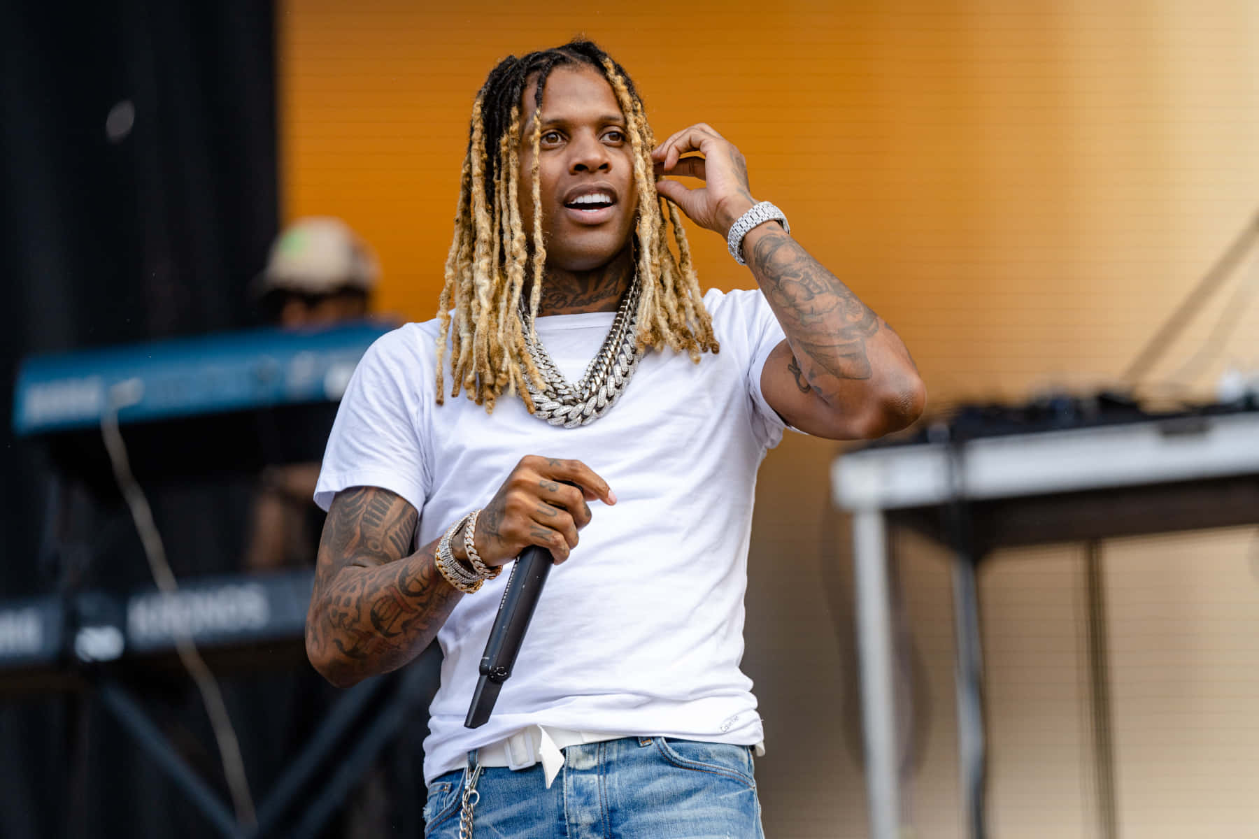 lil durk 桌布 壁紙 — 一位男性饒舌歌手留著金色辮子，手持麥克風，身穿白色T恤和牛仔褲在舞台上表演。