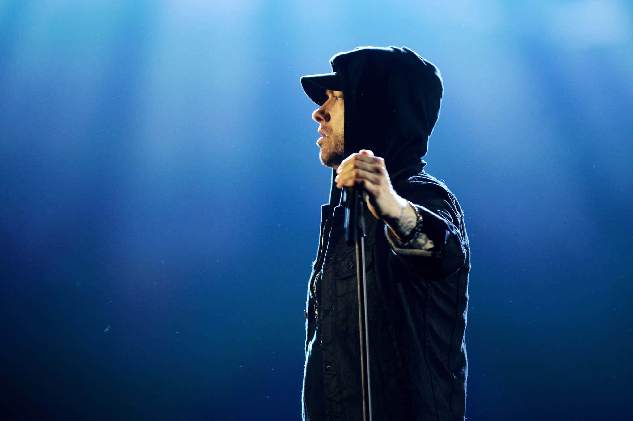 Eminem 桌布 壁紙 — 一位男性饒舌歌手穿著黑色連帽衫和帽子，側身站在戲劇性的藍色舞台燈光下，手持麥克風。