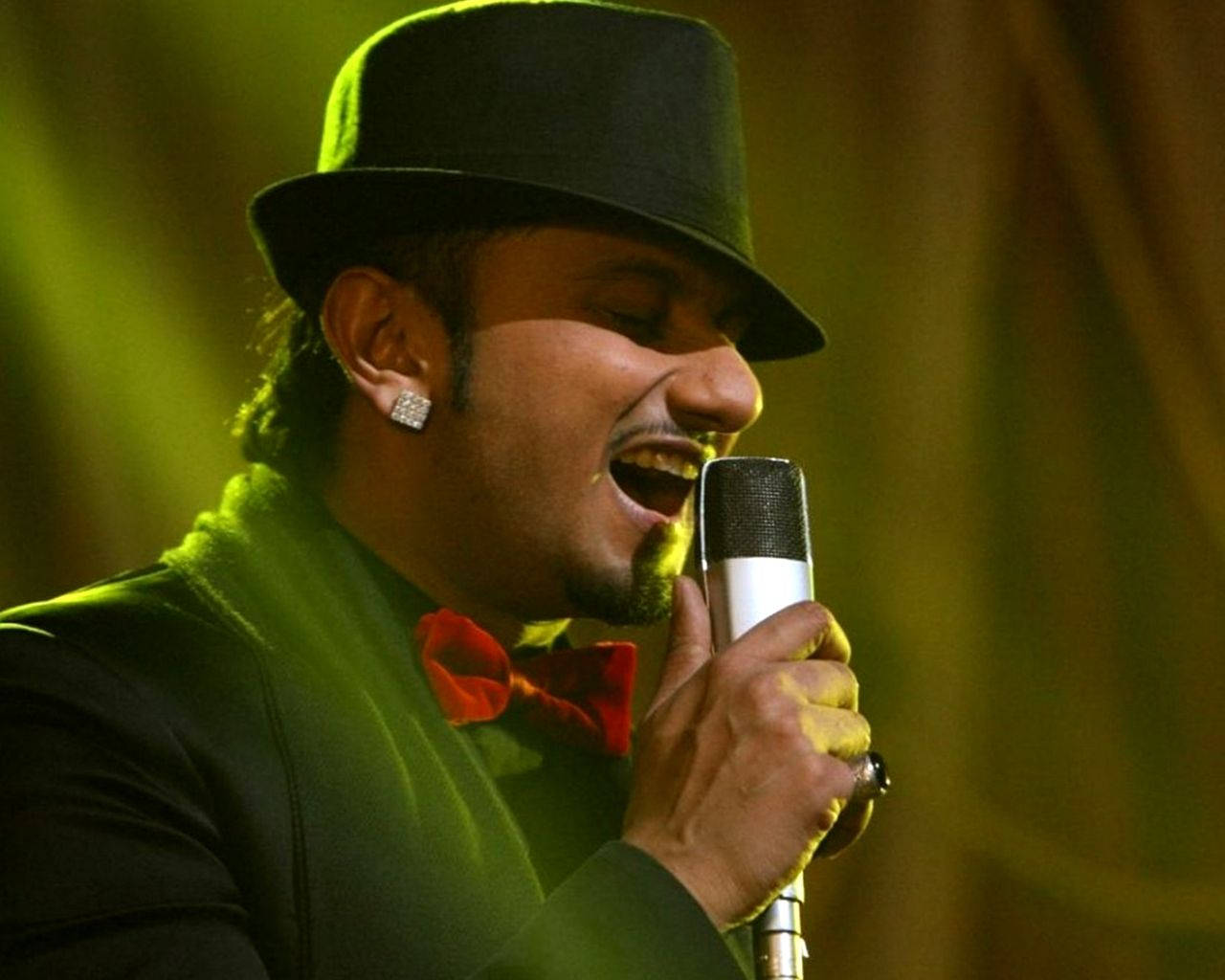Yo Yo Honey Singh《The Xpose》 壁紙 — 一位男歌手戴著黑色高頂帽和紅色領結，在溫暖的舞台燈光下熱情地對著麥克風演唱。