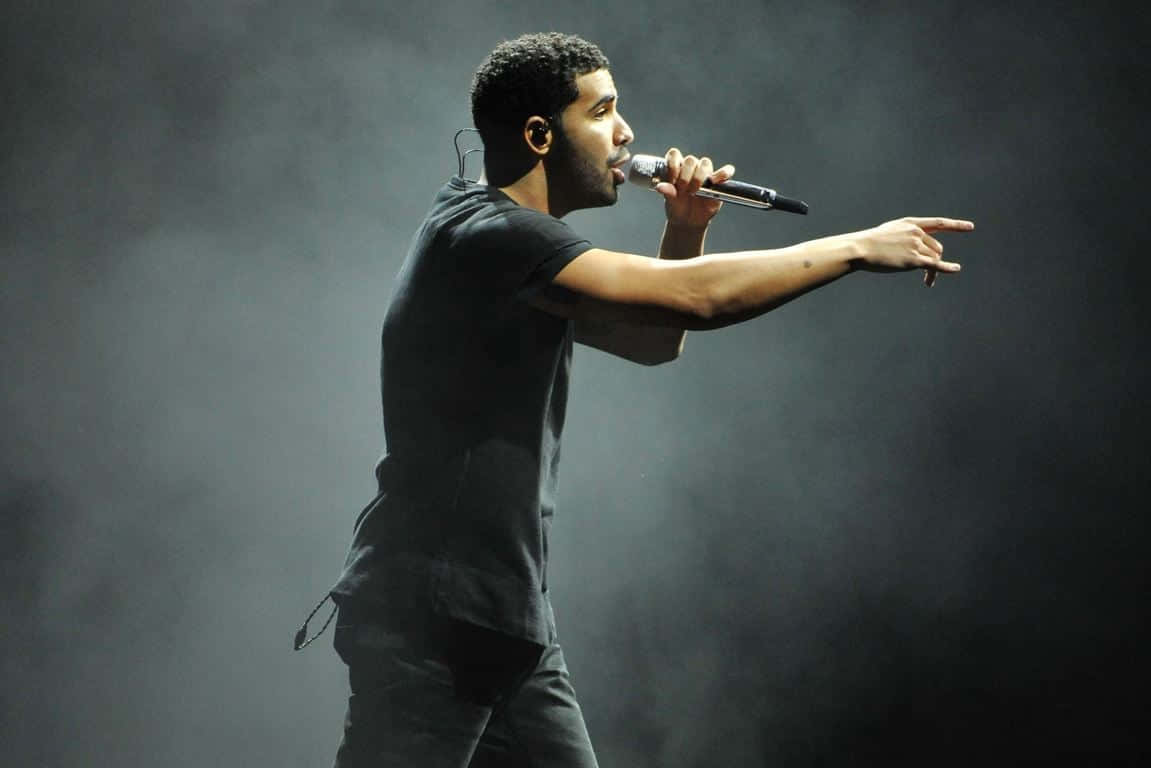 Drake 桌布 壁紙 — 一位男歌手穿著深色衣服在舞台上表演，手持麥克風並在戲劇性的舞台燈光下向前指。