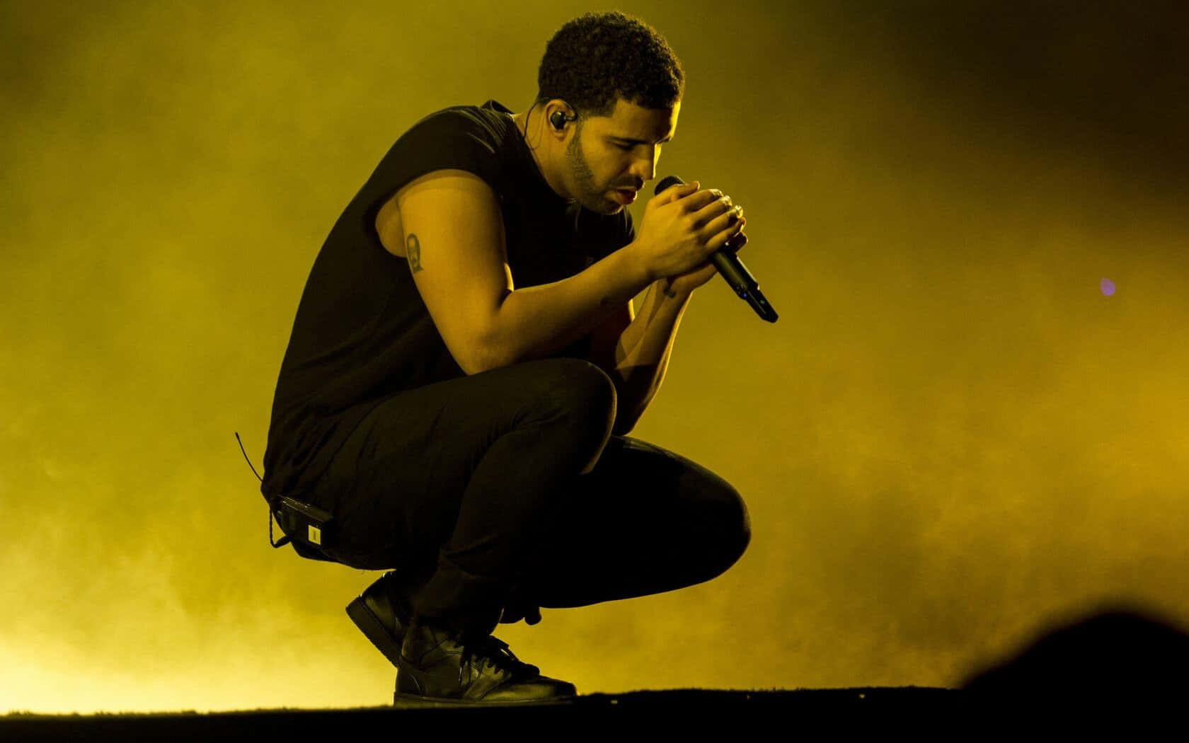 Drake 桌布 壁紙 — 一位男歌手蹲在舞台上，沐浴在金黃色的舞台燈光中，專注地握著麥克風。