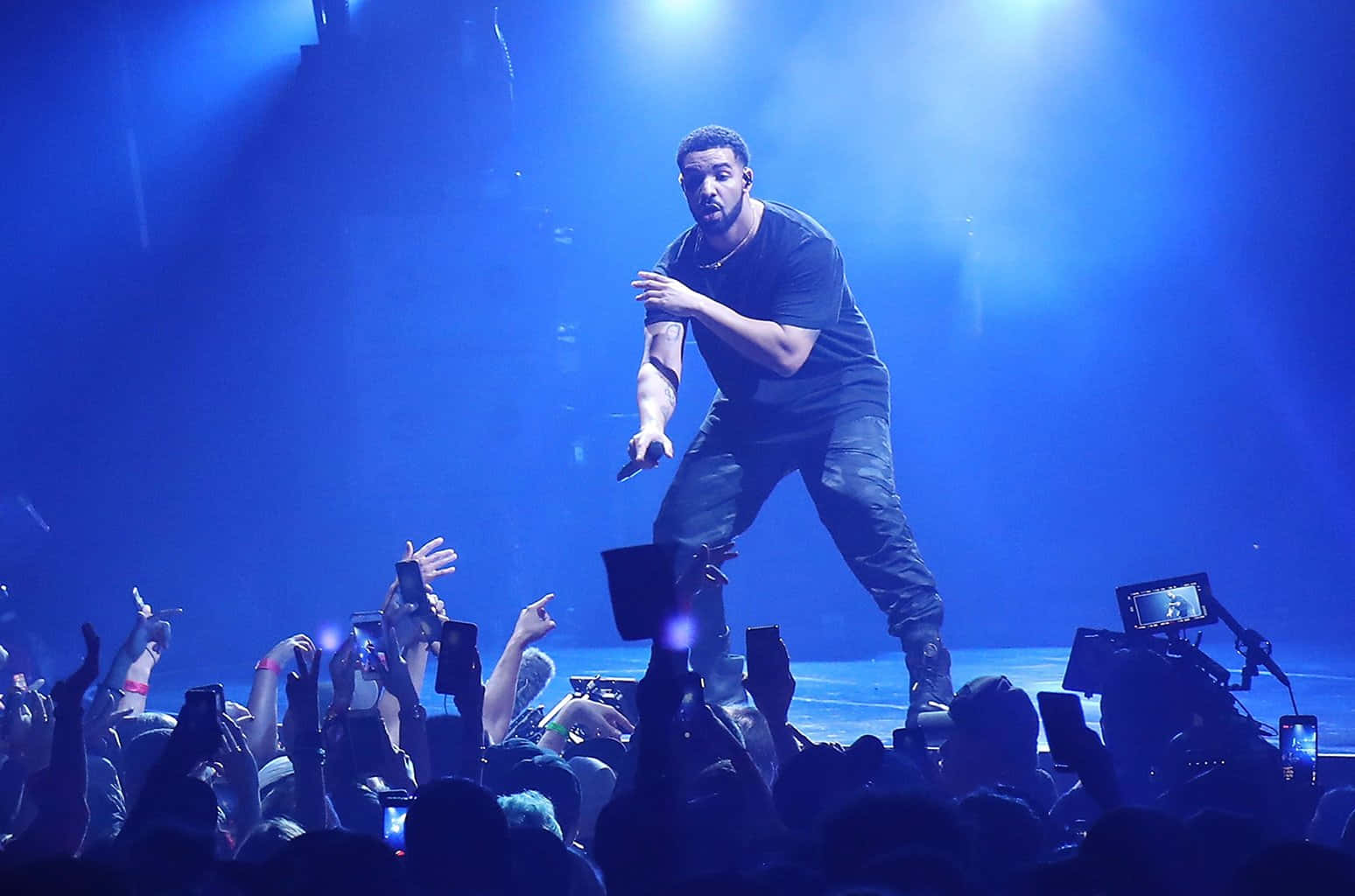 Drake 桌布 壁紙 — 一位男表演者掌控著被強烈藍光籠罩的演唱會舞台，充滿活力的觀眾舉起手機。