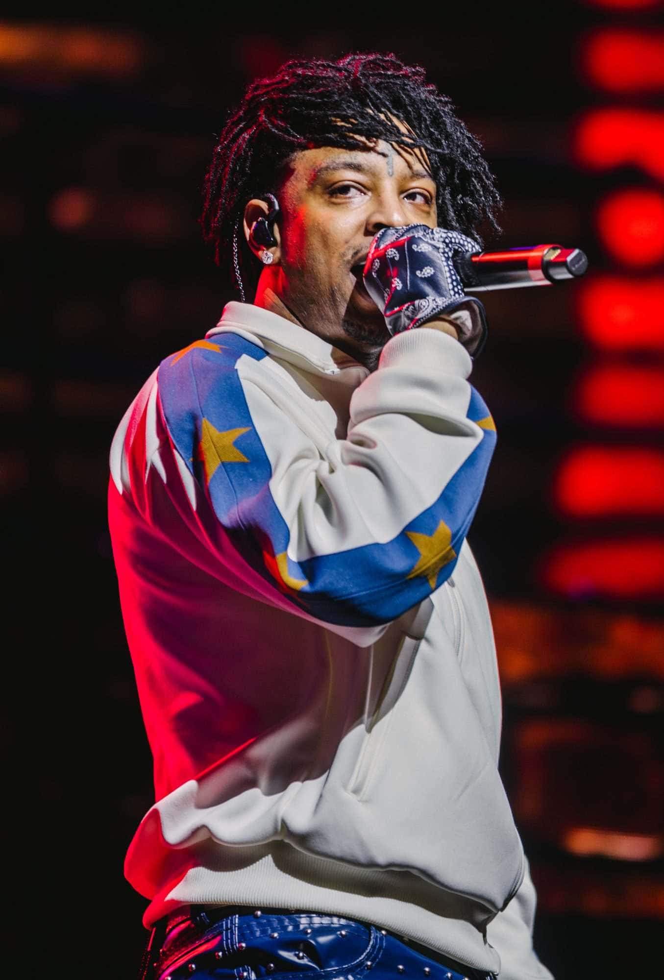 21 Savage 壁紙 — 一位男饒舌歌手梳著辮子，在紅色舞台燈光下，穿著星星圖案連帽衫，對著麥克風熱情表演。