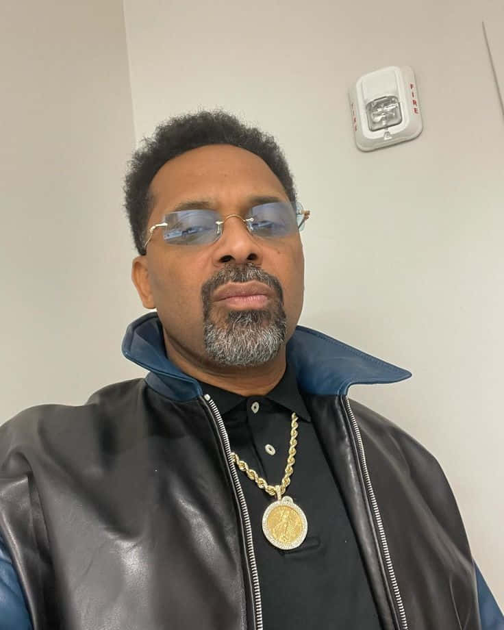 Mike Epps 桌布 壁紙 — 一位留山羊鬍、戴藍色眼鏡的男子，身穿黑色皮褸和金色鏈條，站在有火警警報器的素牆前。