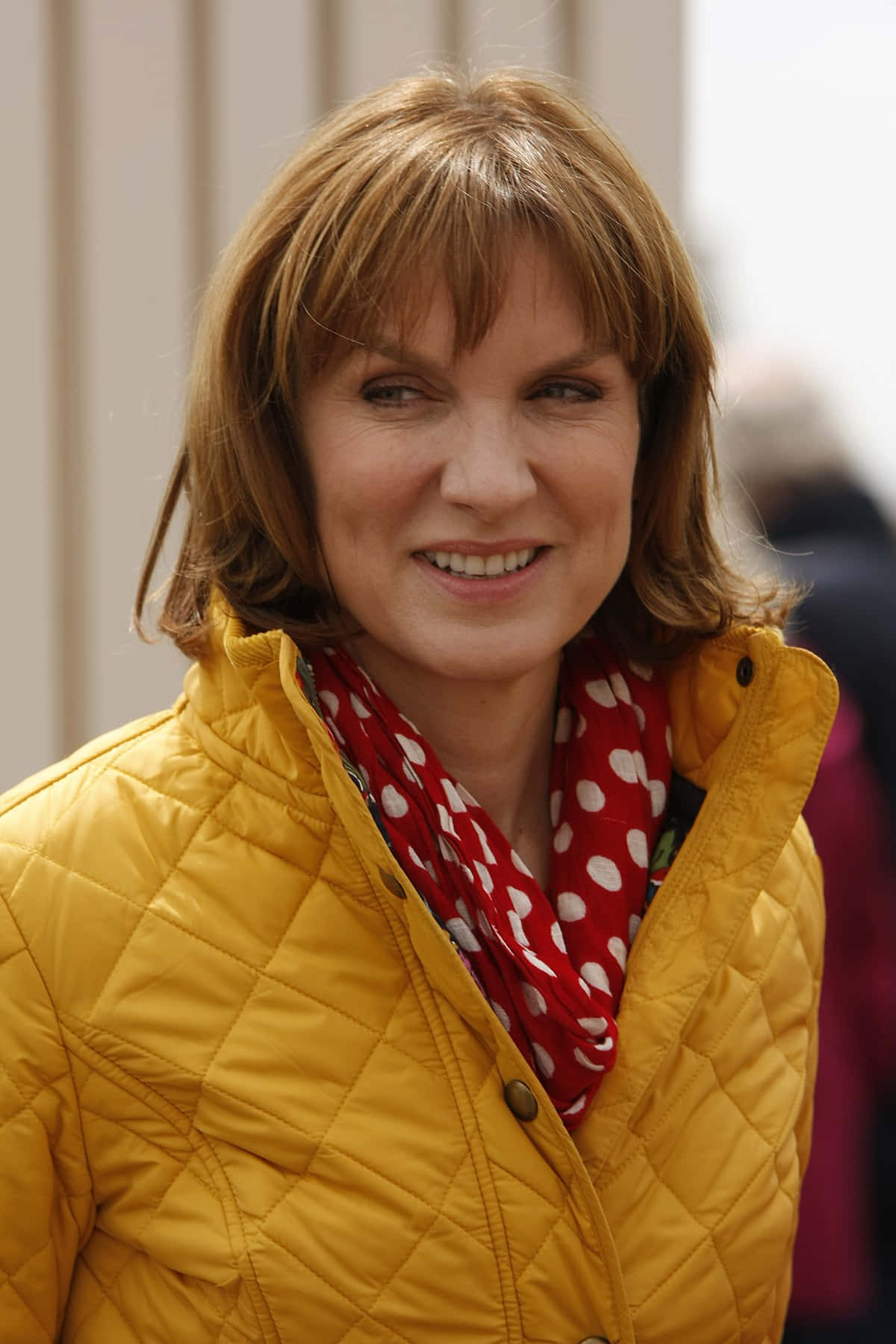 Fiona Bruce 桌布 壁紙 — 一位留著及肩棕色頭髮的女性，身穿亮黃色絎縫外套和紅色圓點圍巾，露出溫暖的笑容。