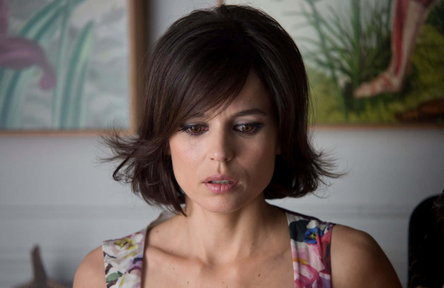 Elena Anaya 桌布 壁紙 — 一位留著深色波波頭、身穿花卉圖案上衣的女性，在柔和光線的室內環境中向下凝視。