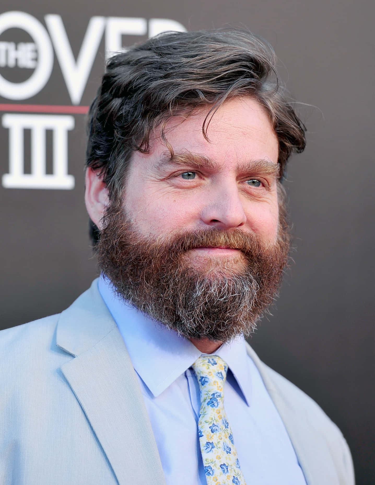 Zach Galifianakis 桌布 壁紙 — 一位留著濃密鬍鬚、藍眼睛的男人，穿著淺灰色西裝和花卉領帶，在電影首映禮上微笑。