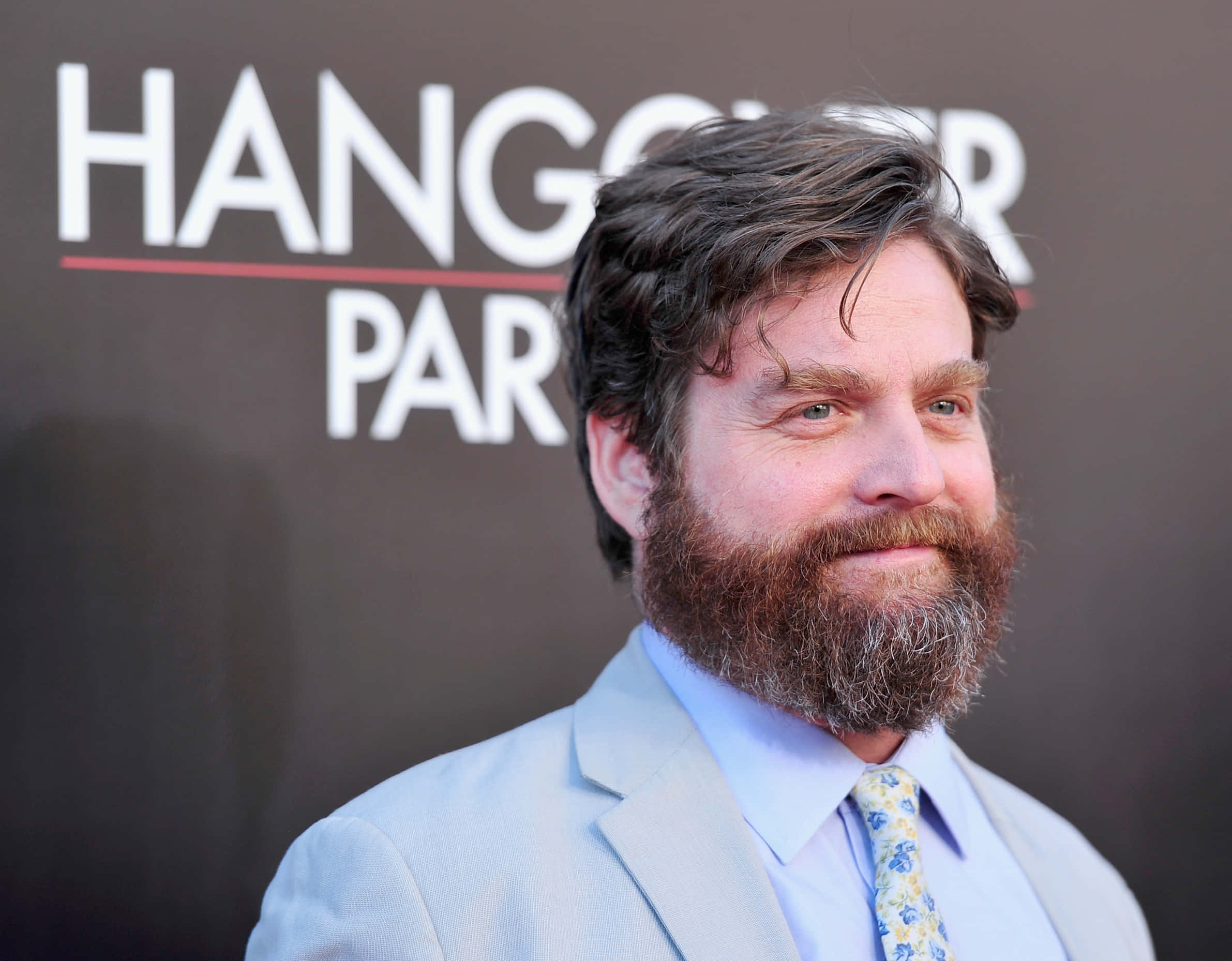 Zach Galifianakis 桌布 壁紙 — 一位留著濃密鬍鬚、頭髮凌亂的男子，在「醉後大丈夫」活動中穿著淺色西裝和圖案領帶對著鏡頭微笑。