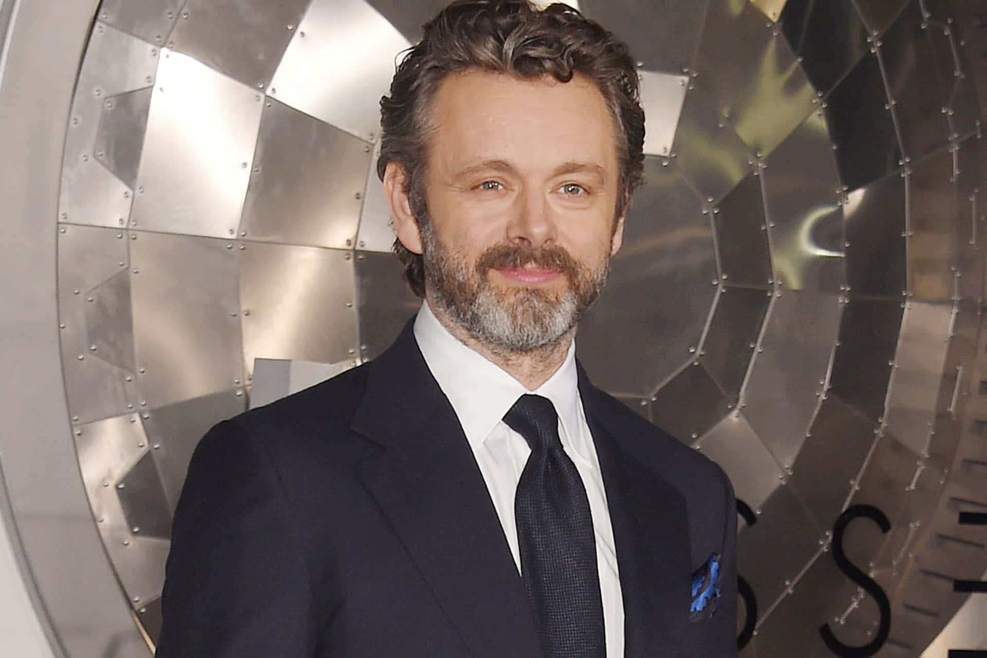 Michael Sheen 桌布 壁紙 — 一位留著灰白鬍鬚、身穿深色西裝的男子自信地站在金屬幾何牆前。