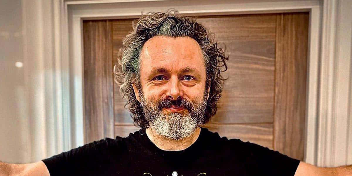 Michael Sheen 桌布 壁紙 — 一位留著灰色捲髮和鬍鬚的男子，在室內穿著黑色T恤，溫暖地微笑。