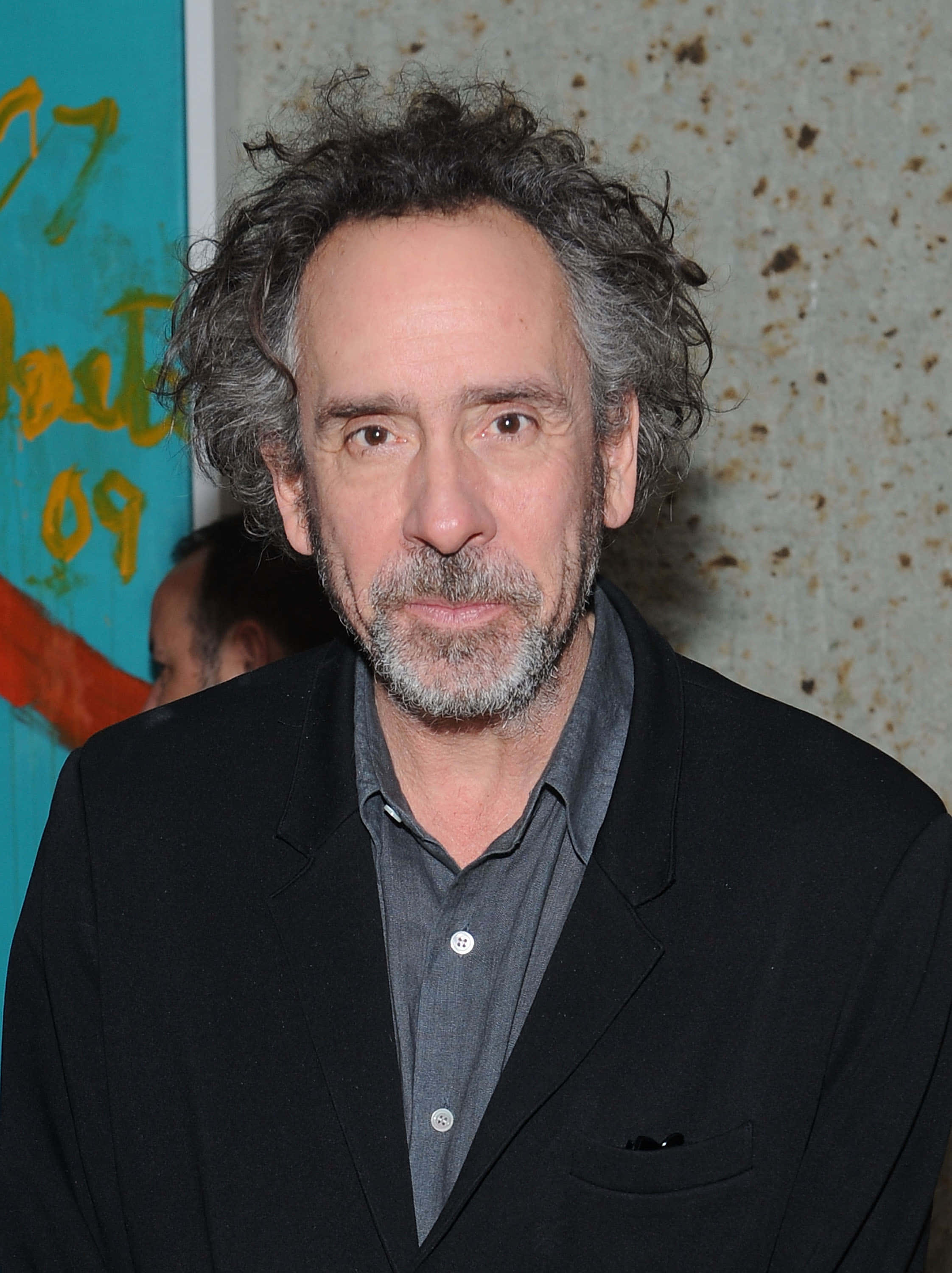 tim burton 桌布 壁紙 — 一位留著灰色捲髮和鬍鬚的男子，身穿黑色西裝外套，內搭灰色襯衫，背景是帶有彩色塗鴉的紋理牆壁。