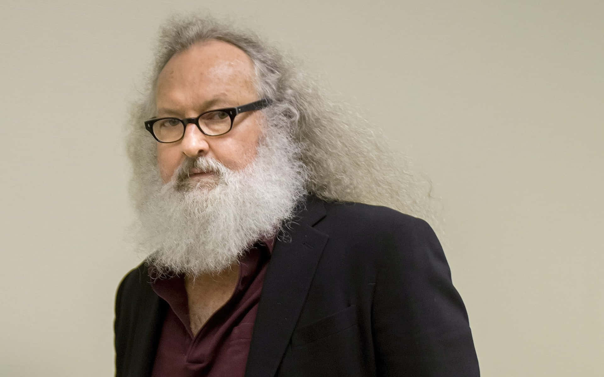 Randy Quaid 桌布 壁紙 — 一位留著灰色長髮和白色濃密鬍鬚的男人，戴著黑框眼鏡，身穿深色西裝外套，內搭栗色襯衫，背景是純米色牆壁。