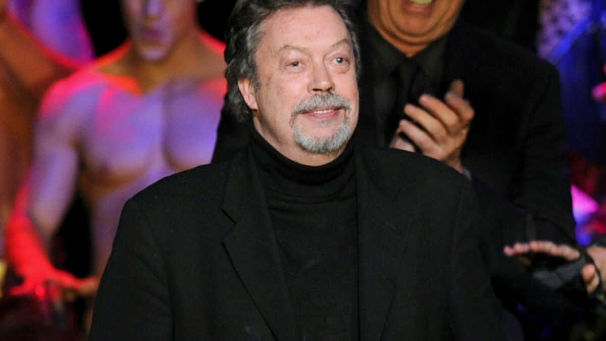 Tim Curry 桌布 壁紙 — 一位留著灰色鬍鬚和鬍子的男士，身穿黑色高領毛衣和西裝外套，在一個色彩繽紛的活動中對著鏡頭微笑，身後有模糊的人影。