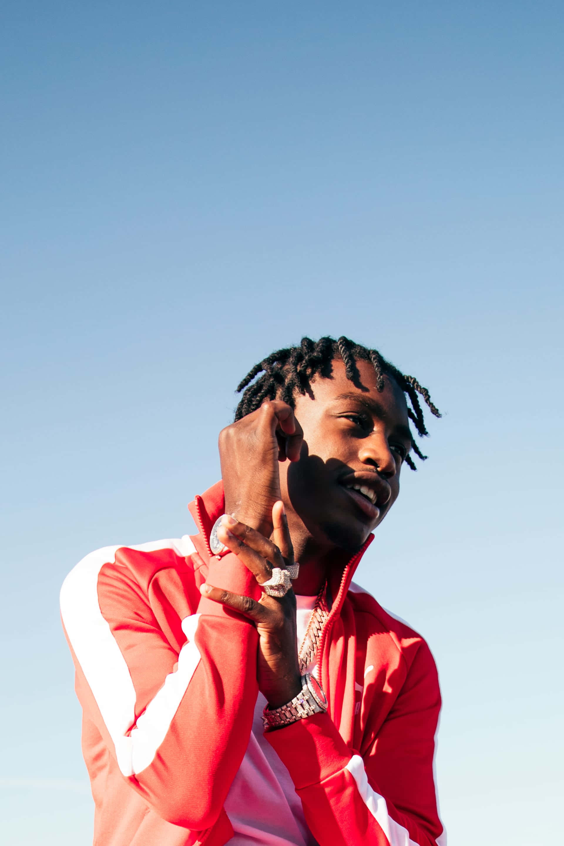 Lil Tjay 桌布 壁紙 — 一位留著辮子、身穿紅色運動外套的年輕男子，在晴朗的藍天下微笑著調整耳機。