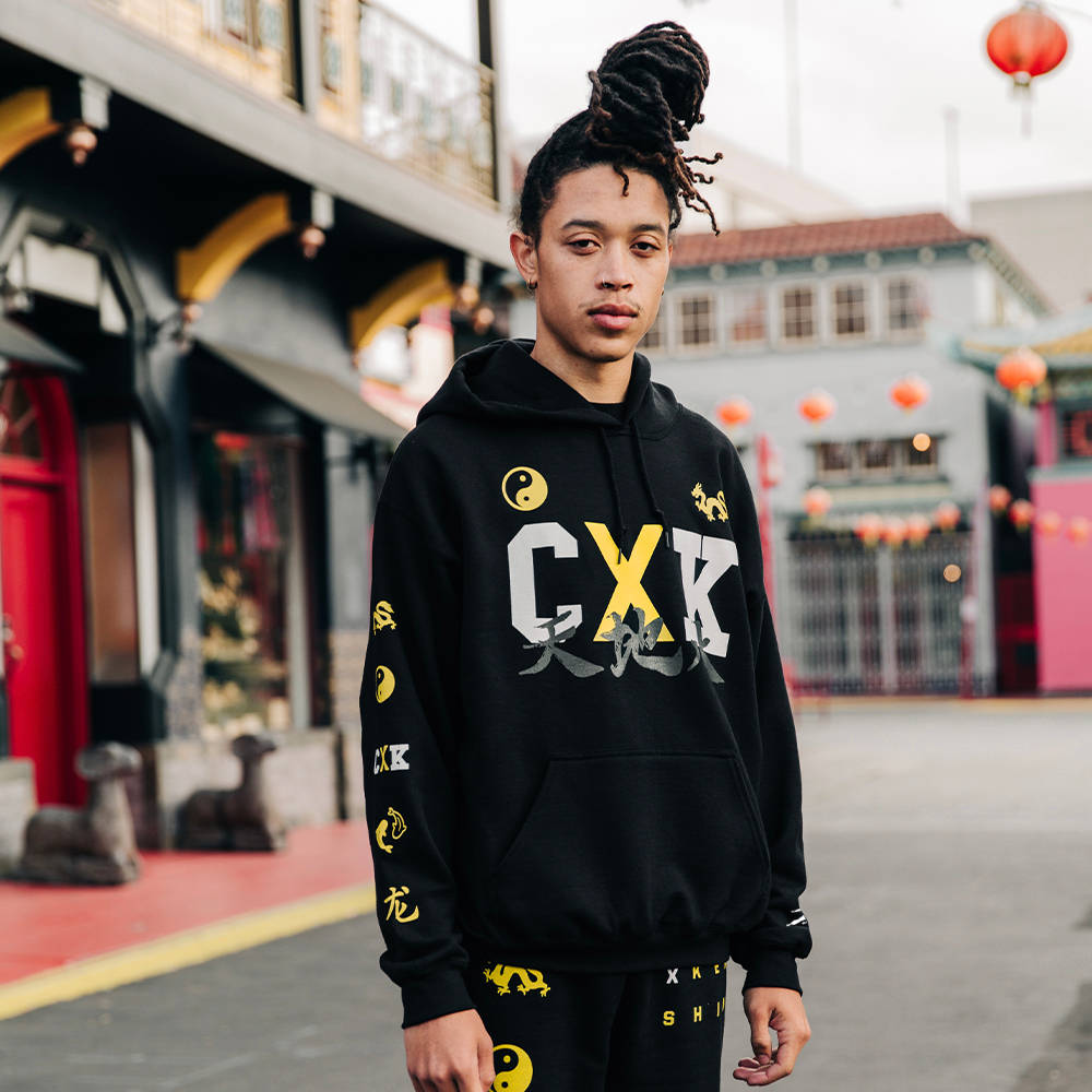 Coryxkenshin 桌布 壁紙 — 一位留著辮子的年輕人自信地站在唐人街的街道上，身穿帶有醒目黃白圖案的黑色連帽衫。