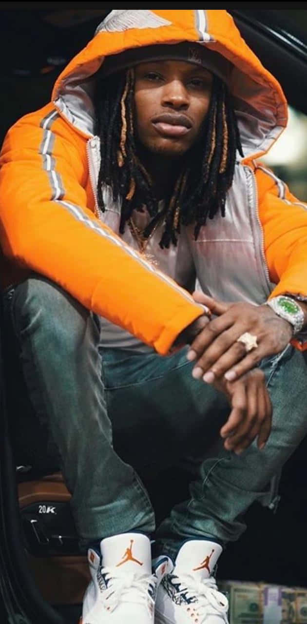 King Von 桌布 壁紙 — 一位留著辮子的年輕男子自信地坐在車裡，身穿亮橙色羽絨外套和白色Jordan運動鞋。