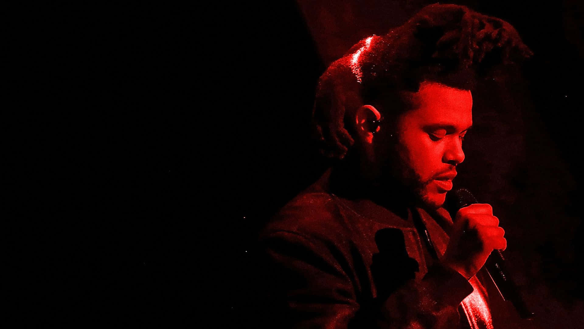 The Weeknd 桌布 壁紙 — 一位留著辮子的男歌手在黑暗場地中，於強烈的紅色舞台燈光下拿著麥克風表演。