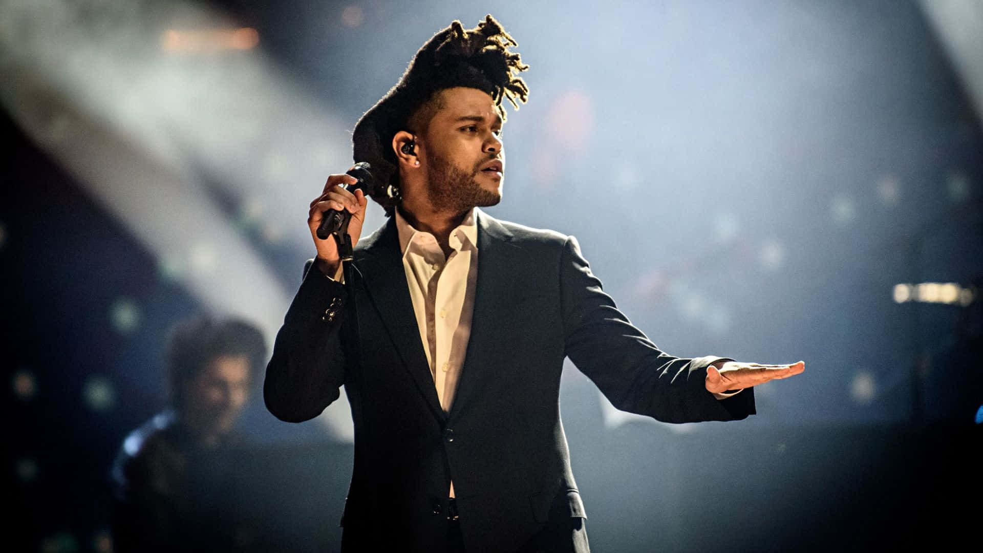 The Weeknd 桌布 壁紙 — 一位留著辮子的男歌手穿著深色西裝，在戲劇性的聚光燈和煙霧效果下在舞台上表演。