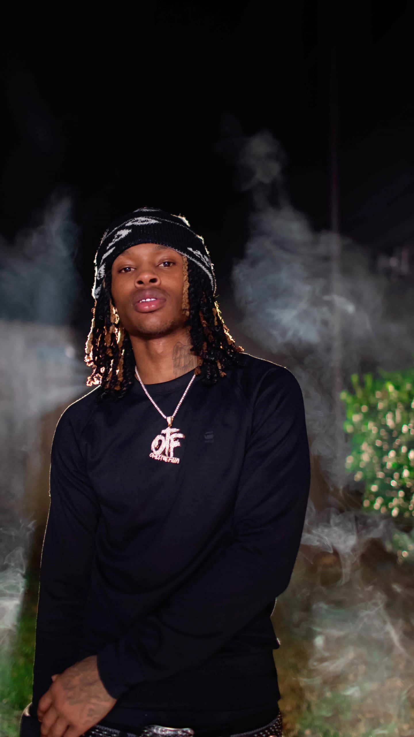 King Von 桌布 壁紙 — 一位留著辮子頭、戴黑色冷帽的年輕男子，自信地站在煙霧瀰漫的黑暗中，身穿帶有粉紅色標誌的黑色長袖上衣。