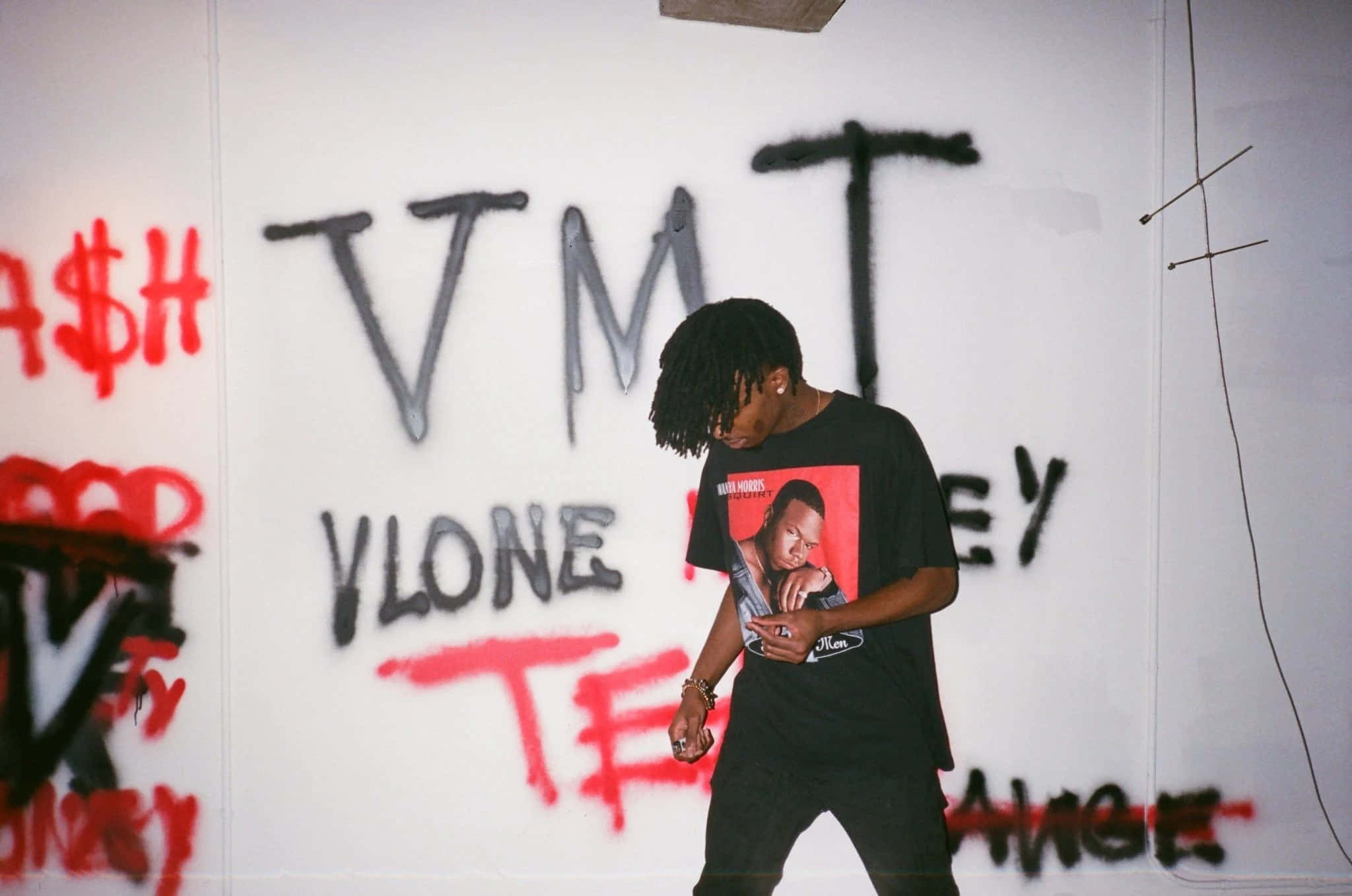 Vlone 桌布 壁紙 — 一位留著辮子頭的年輕男子站在塗鴉牆前，身穿帶有紅色圖案的黑色T恤。
